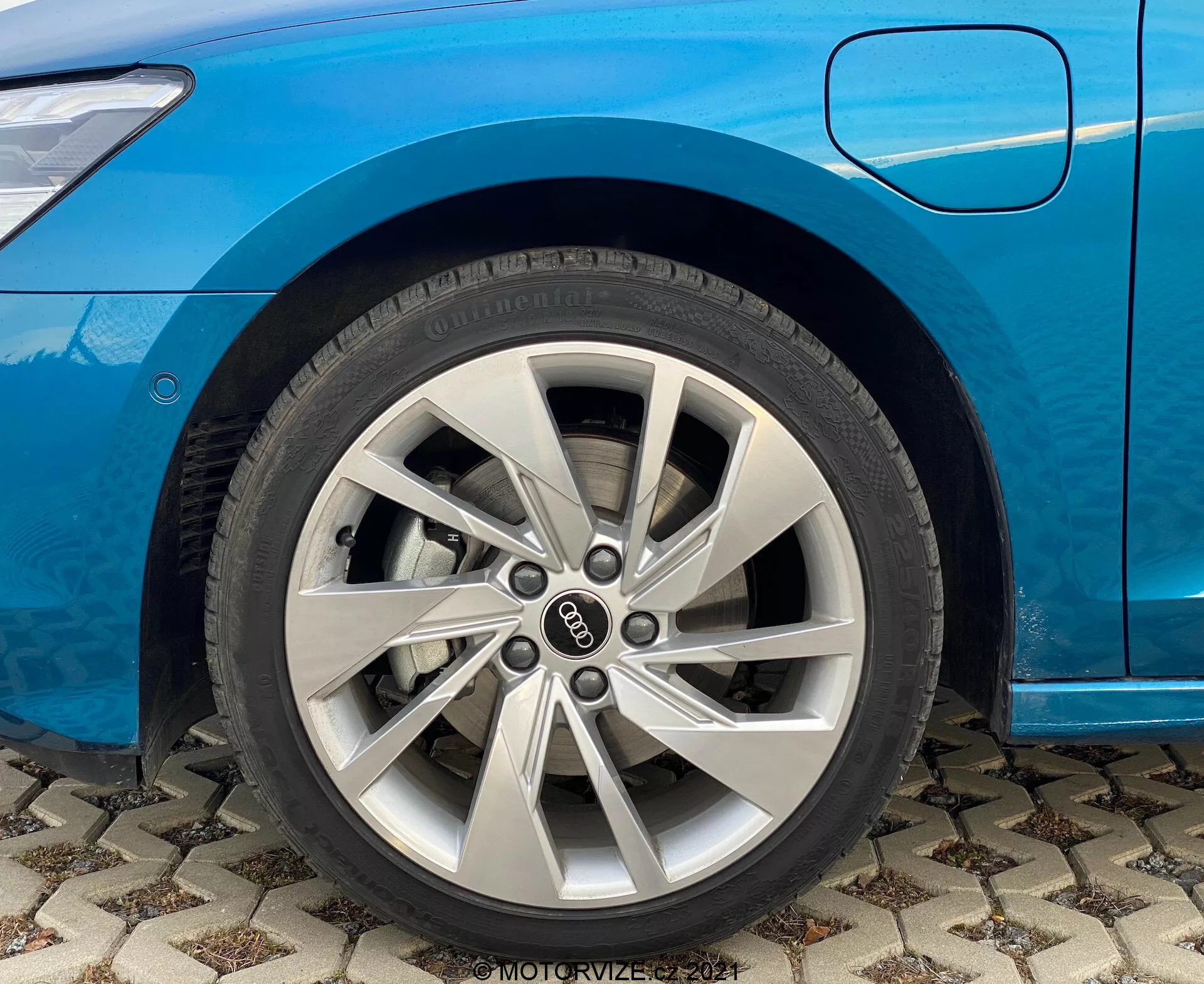 Detailní pohled na přední kolo modré Audi A3 2020 (A3 Sportback). Zaměřeno na stříbrné vícepaprskové hliníkové kolo na straně spolujezdce, viditelný přední blatník a spodní okraj dveří spolujezdce. Auto je zaparkované na vzorované dlažbě a je vidět vzorek pneumatik spolu s brandingovými detaily. Významné pro diskuze o automobilovém designu a výrobních technikách kol. Obsahuje vodoznak 'MOTORVIZE 2021.'
