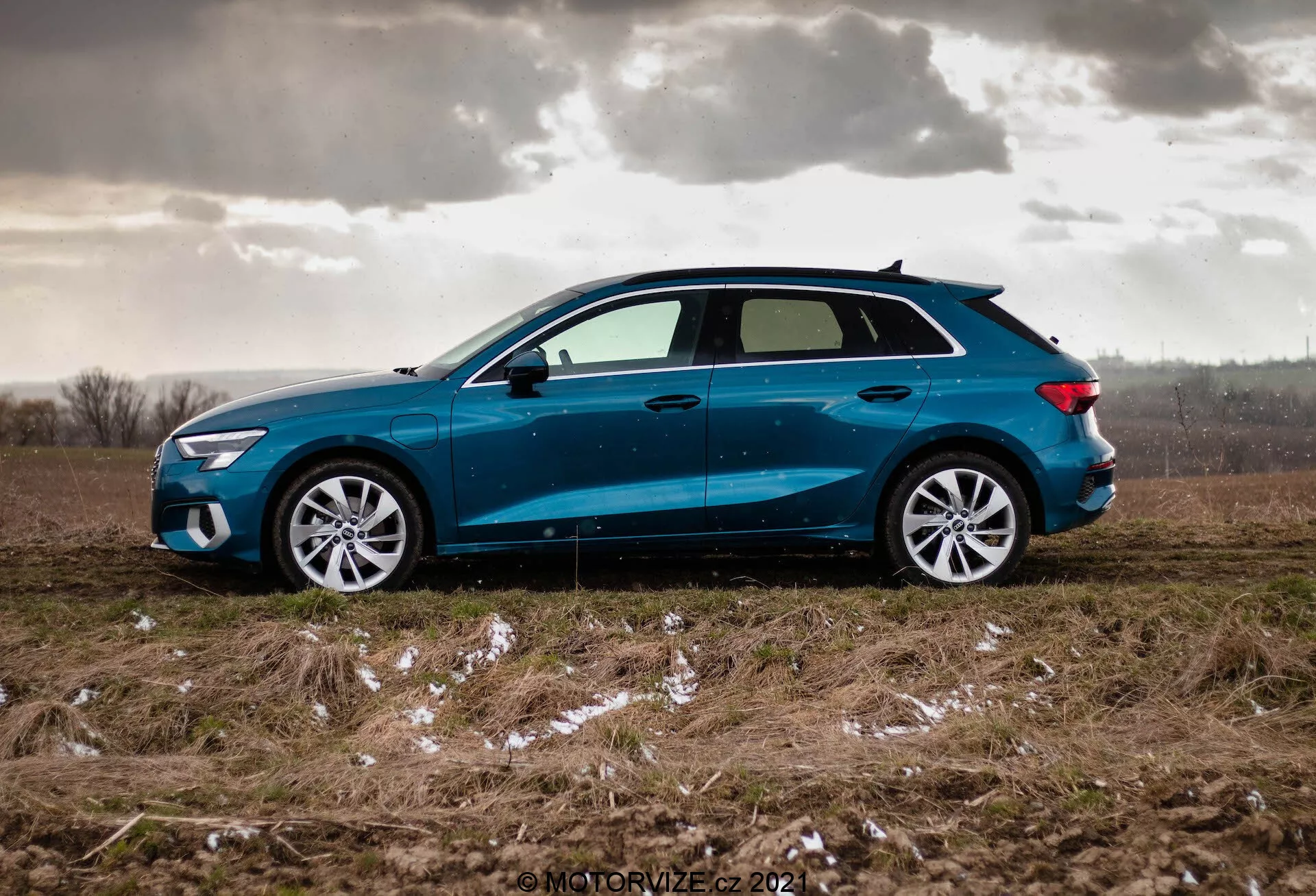 Boční pohled na modrý Audi A3 Sportback z roku 2020 zaparkovaný na nerovném travnatém poli s náznaky sněhu nebo ledu a zataženou oblohou, zdůrazňující dynamický design vozu s mírně skloněnou střechou, vícelopatkovými hliníkovými ráfky, lehce zabarvenými okny, bočními zrcátky v barvě karoserie s integrovanými směrovými světly a ostrými LED světlomety a zadními světly.