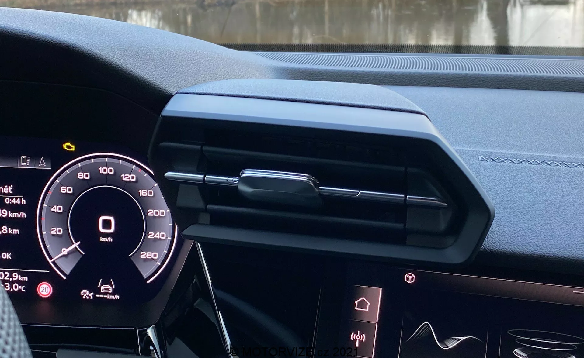 Pohled na interiér modelu Audi A3 Sportback 2020 z místa řidiče, zobrazující digitální přístrojovou desku, elegantní design palubní desky s uzavřeným centrálním větrákem a dekorativním obložením, okraj infotainmentového displeje, luxusní kožené materiály s prošitím, ambientní osvětlení a částečně viditelný volant s možnými ovládacími prvky, zdůrazňující moderní automobilovou technologii a luxusní design.