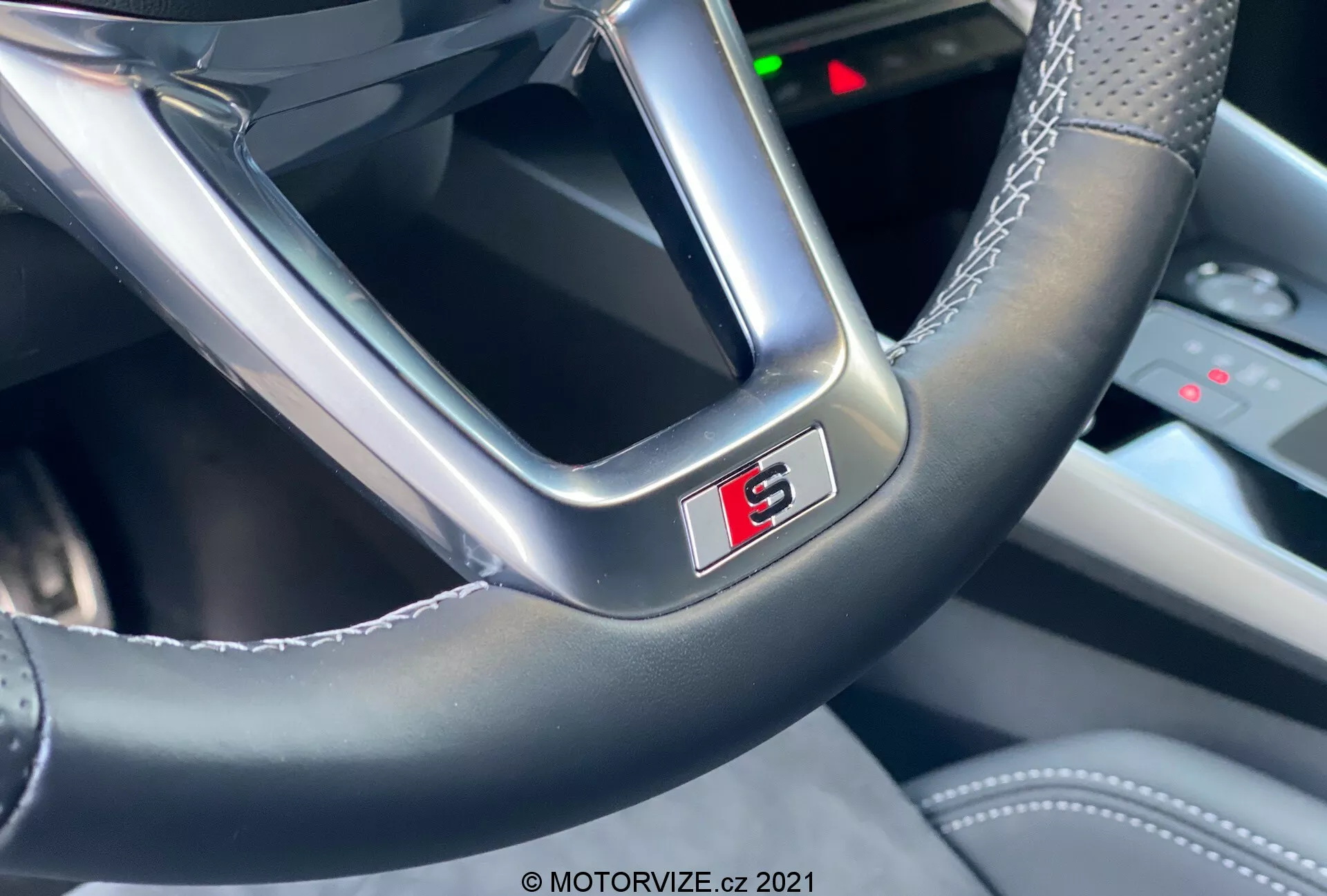 Detailní pohled na volant moderního auta z pohledu řidiče, zaměřený na spodní část s výrazným 'S' odznakem lemovaným červenou. Volant ukazuje kombinaci hladké kůže a lesklejšího materiálu s viditelným prošíváním, zdůrazňující řemeslné zpracování a sportovní charakter Audi A3 2020 Sportback.