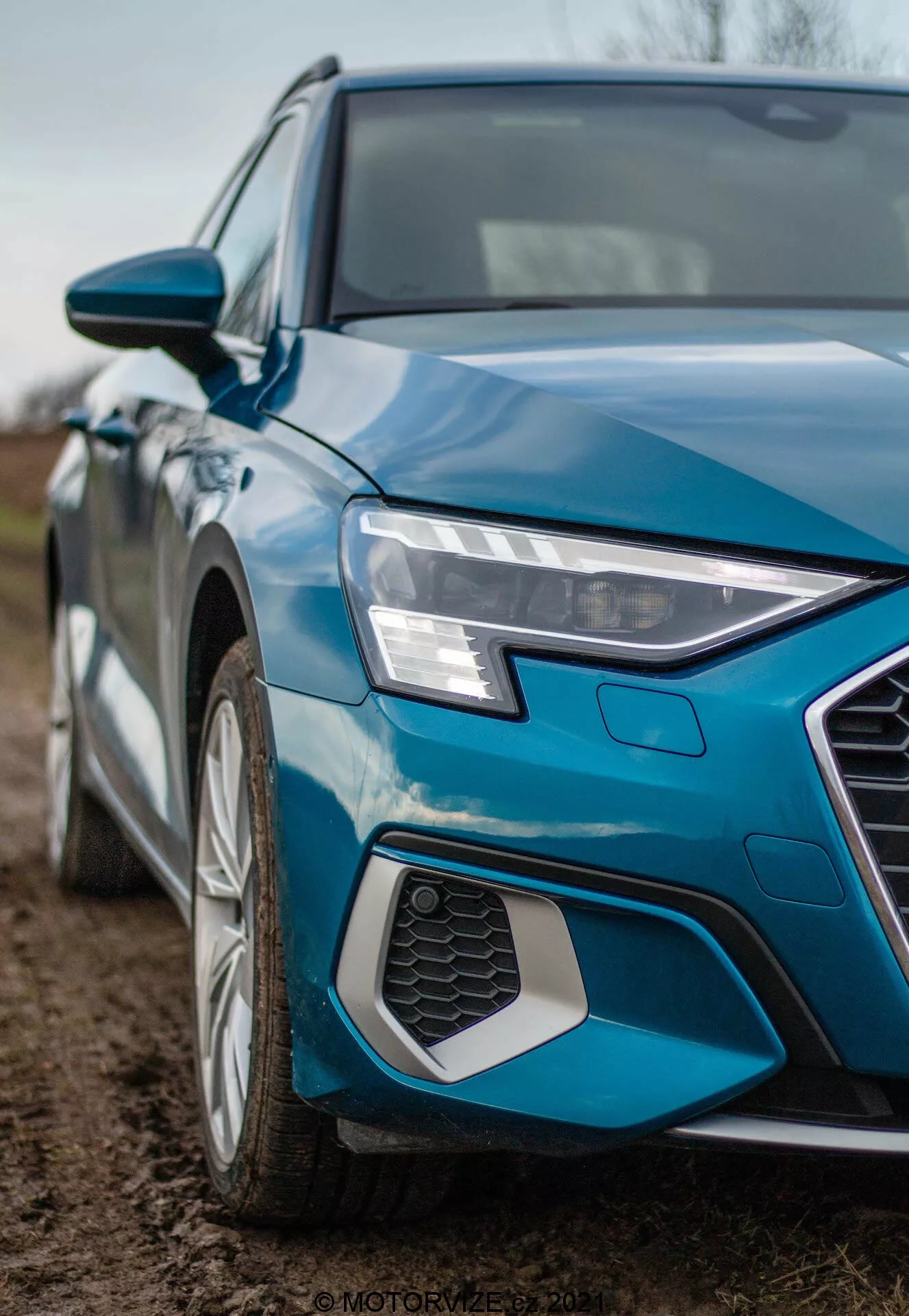 Detailní snímek Audi A3 Sportback 2020 z úhlopříčného pohledu, zaměřený na levý přední kvadrant. Fotografie z nízkého úhlu zachycuje ostré a hranaté přední levé světlomety s LED denním svícením, část šestiúhelníkové mřížky s voštinovým vzorem, levé přední blatník a část dveří řidiče. Také jsou vidět vícepaprsková hliníková kola, metalizovaná modrá barva a zrcátko v barvě karoserie s integrovanými směrovkami, zdůrazňující moderní design a technologické prvky.