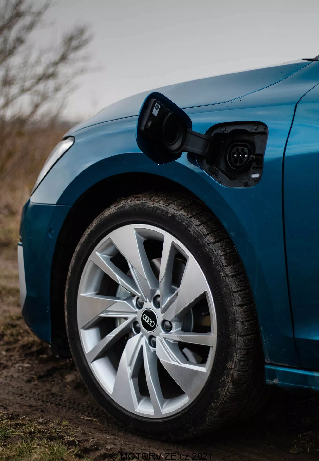 Detailní záběr na přední část řidičské strany modrého Audi A3 2020 Sportback, zdůrazňující kolo a nabíjecí port. Obrázek, pořízený z nízkého úhlu směrem k přední části, ukazuje otevřený nabíjecí port nad předním kolem s připojeným nabíjecím kabelem. Mělká hloubka ostrosti ostře zaostřuje na kolo a nabíjecí port, odhaluje typické překládající se kruhy loga Audi a vícepaprskový design kola s nízkoprofilovou pneumatikou, vše v mírně tlumeném, možná soumrakovém osvětlení.