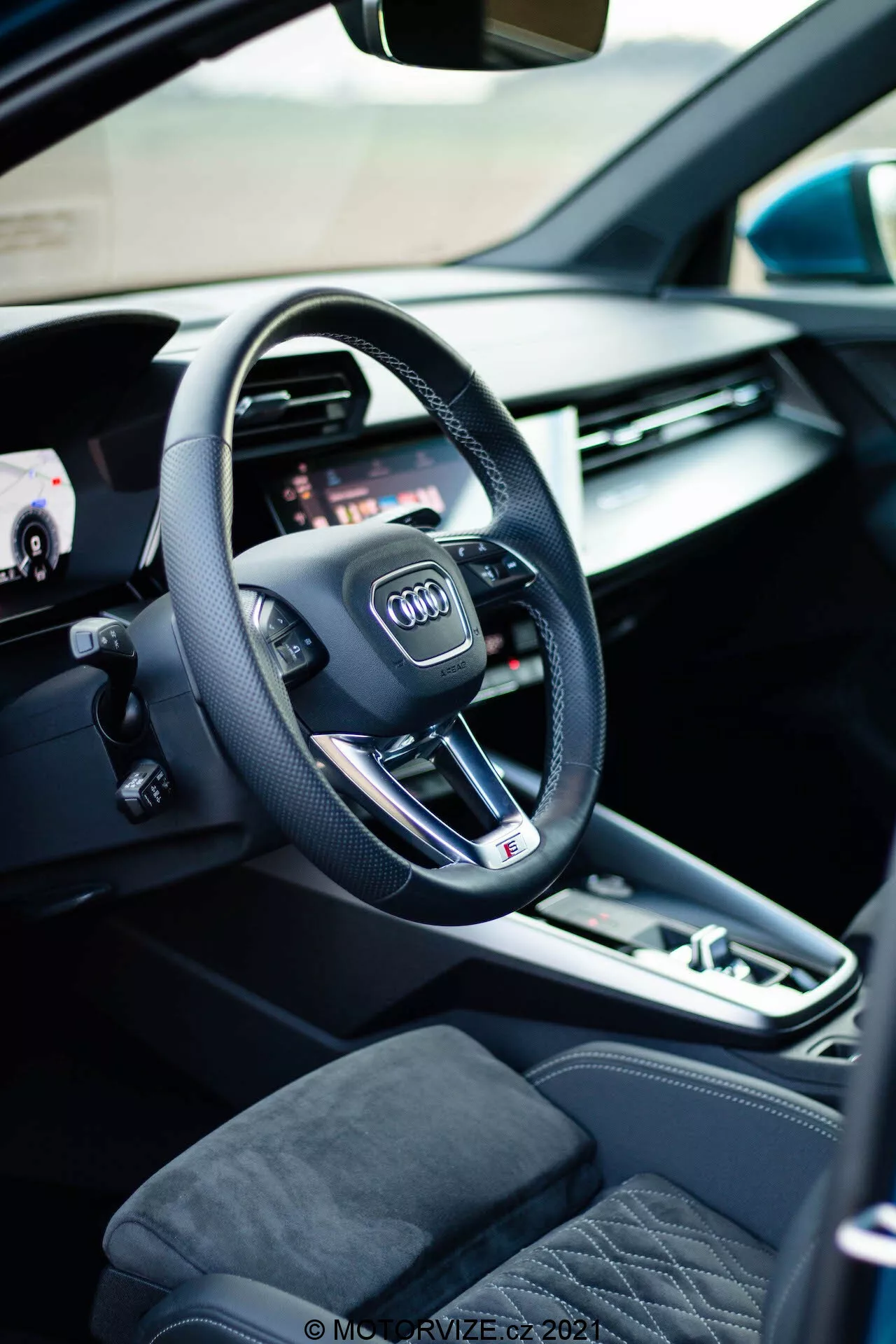 Pohled na interiér Audi A3 Sportback 2020 z pohledu řidiče, kde je vidět moderní tříramenný volant s logem Audi, integrovanými multifunkčními tlačítky a plochým spodkem, což naznačuje sportovní výkon. Za volantem je vidět část digitálního přístrojového panelu nebo virtuálního kokpitu, který nabízí přizpůsobitelné zobrazení pro různé funkce vozu. Vlevo, otevřené dveře ukazují ovládání elektrických oken a paměťové nastavení sedadel, zdůrazňující pohodlí a přizpůsobení. Středová konzola zahrnuje automatický volič převodovky, tlačítko elektronické parkovací brzdy a dotykovou podložku/ovládací kolečko pro infotainment systém, zdůrazňující zapojení řidiče. Viditelná sedadla jsou čalouněna prémiovými materiály s prošitým designem, což přidává na luxusním pocitu z interiéru.