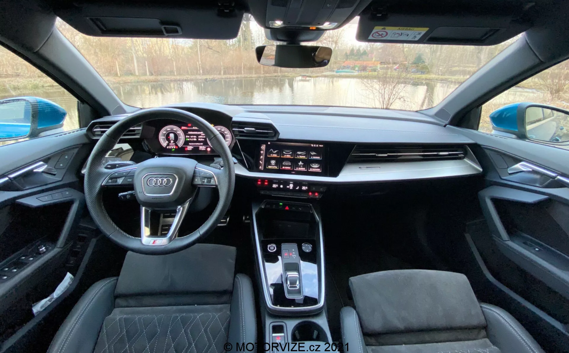 Interiér modelu Audi A3 Sportback 2020 z pohledu řidiče, zobrazující moderní palubní desku s digitálním přístrojovým panelem, sportovním multifunkčním volantem s logem Audi, integrovaným infotainment dotykovým displejem, elegantním středovým panelem s automatickým voličem převodovky, ovládáním klimatu a sportovními sedadly s diamantovým prošíváním pod panoramatickým střešním oknem.