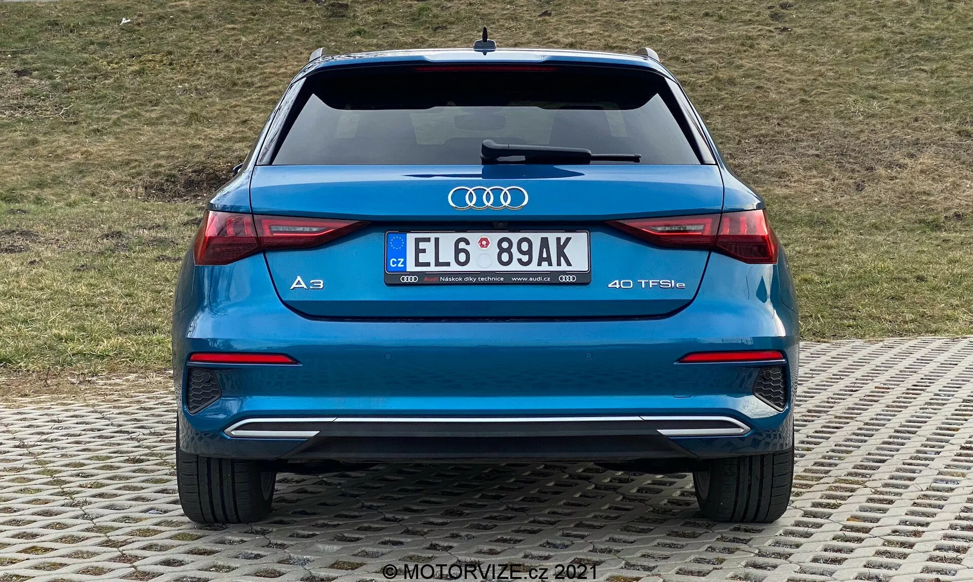 Zadní pohled na modrý Audi A3 2020 Sportback ukazující zadní nárazník, zavazadlový prostor, zadní světla, výfuky a zadní okno. Výrazné logo Audi a označení modelu 'A3' s odznakem '40 TFSI' indikujícím turbo benzínový motor. LED zadní světla s elegantním horizontálním designem, dvojité výfuky, zadní nárazník s difuzorem a parkovacími senzory a evropská SPZ. V pozadí travnatý svah.