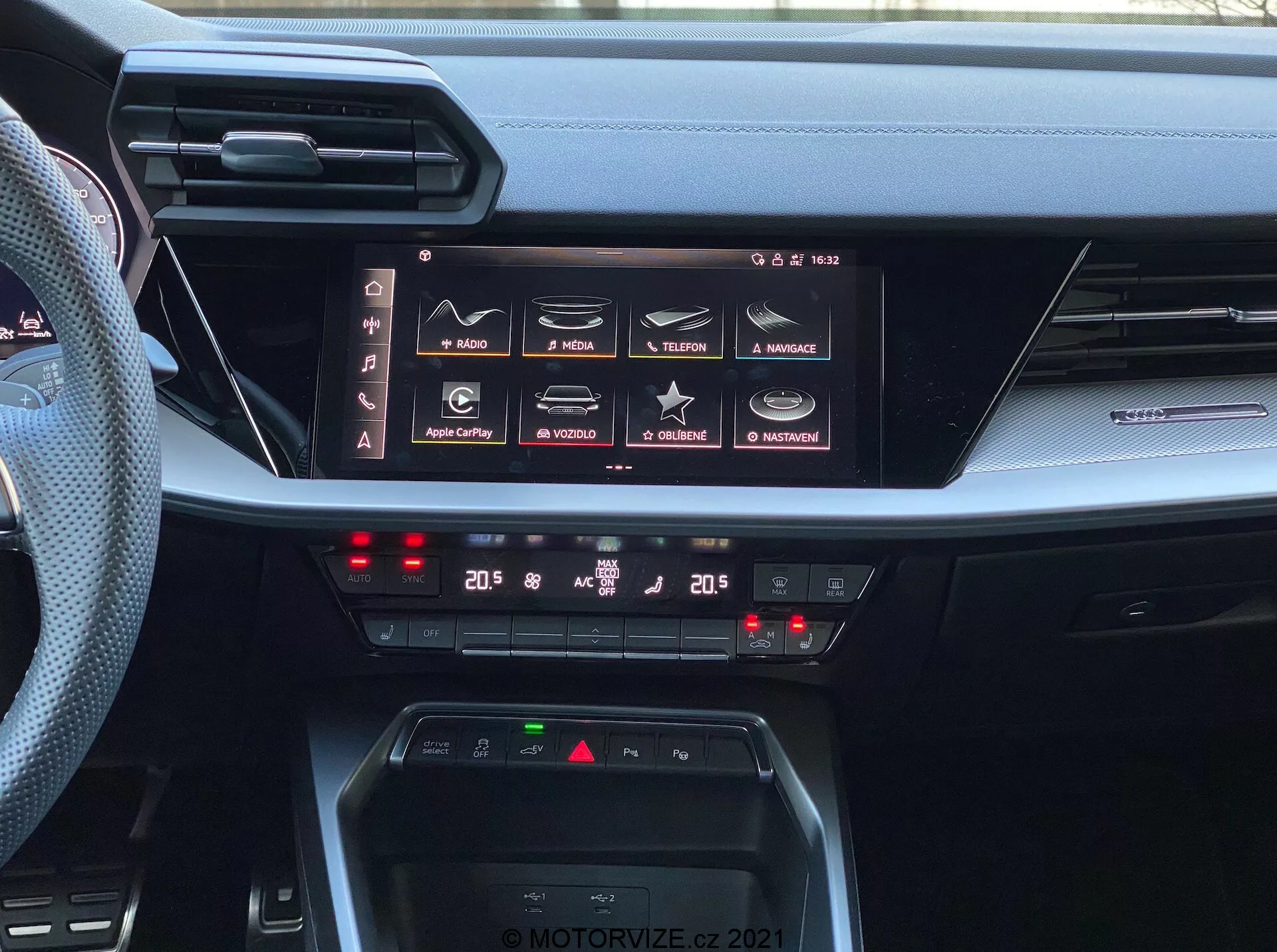 Pohled na interiér palubní desky modelu Audi A3 Sportback 2020, zdůrazňující jeho pokročilý infotainment systém a ovládání klimatizace. Obrázek zachycuje velký horizontální dotykový displej s funkcemi jako navigace, média, telefonní konektivita a nastavení vozidla, naznačující komplexní schopnosti infotainmentu včetně Apple CarPlay a Android Auto. Pod displejem jsou dvojzónové digitální ovladače klimatizace s nastavením na 20 stupňů Celsia pro řidiče i spolujezdce. Palubní deska také zahrnuje oblast pro bezdrátové nabíjení a tenké horizontální větrací otvory, které ilustrují moderní design automobilových interiérů. Foto je opatřeno vodoznakem 'MOTORVIZE.CZ 2021'.