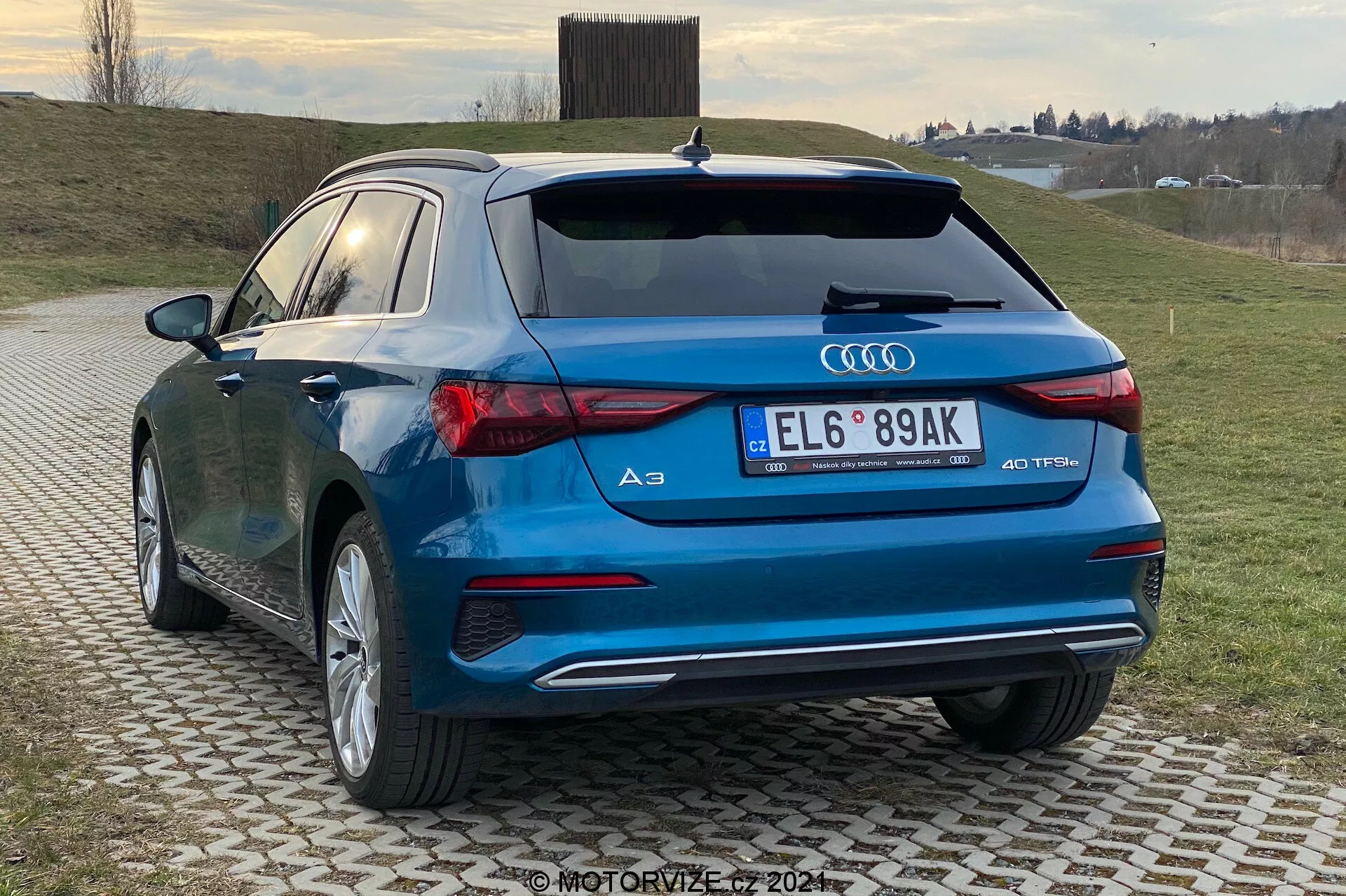 Obrázek zachycuje moderní hatchback, Audi A3 Sportback 2020, ze zadního tříčtvrtečního pohledu, zvýrazňující zadní a boční profil vozidla. Viditelné jsou charakteristické designové prvky, jako LED zadní světla, dvojité výfukové koncovky, emblém Audi kroužků a značka '40 TFSI', což naznačuje sportovní variantu S-line. Auto s lesklým modrým lakem je zaparkováno na vzorované dlažbě v venkovském nebo polo-venkovském prostředí s travnatým pozadím a zataženou oblohou.