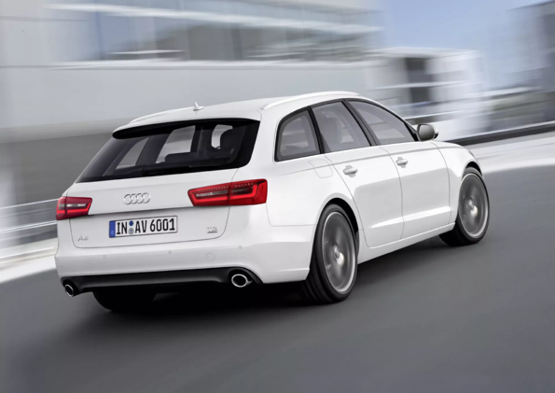 Dynamický pohled zezadu a z boku na bílé kombi Audi A6 Avant 2011 v pohybu, s rozostřenými koly naznačujícími rychlost, dvojitým výfukem, slitinovými koly s viditelnými diskovými brzdami a spoilerem na střeše pro lepší aerodynamiku, snímek pořízen venku.