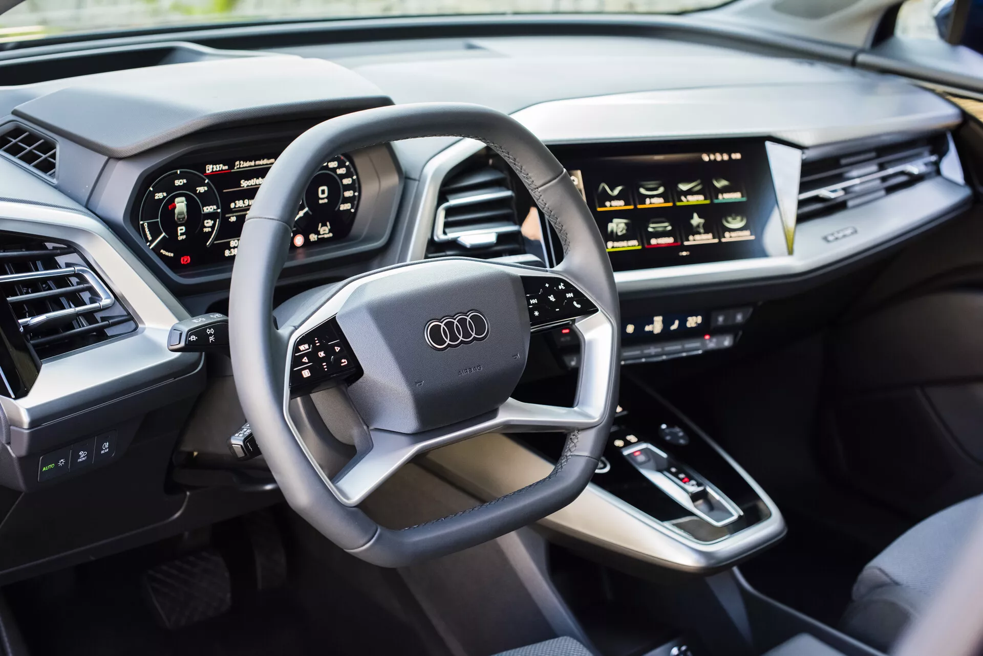 Pohled do interiéru Audi Q4 e-tron 2021 ze strany řidiče s multifunkčním volantem s logem Audi, digitálním přístrojovým panelem, středovou konzolou s řadicí pákou, dotykovými ovladači HVAC, displejem infotainmentu, elegantní palubní deskou s integrovanými výdechy vzduchu a luxusně čalouněnými dveřmi, předvádějící moderní technologie automobilů a prvky luxusních vozidel.