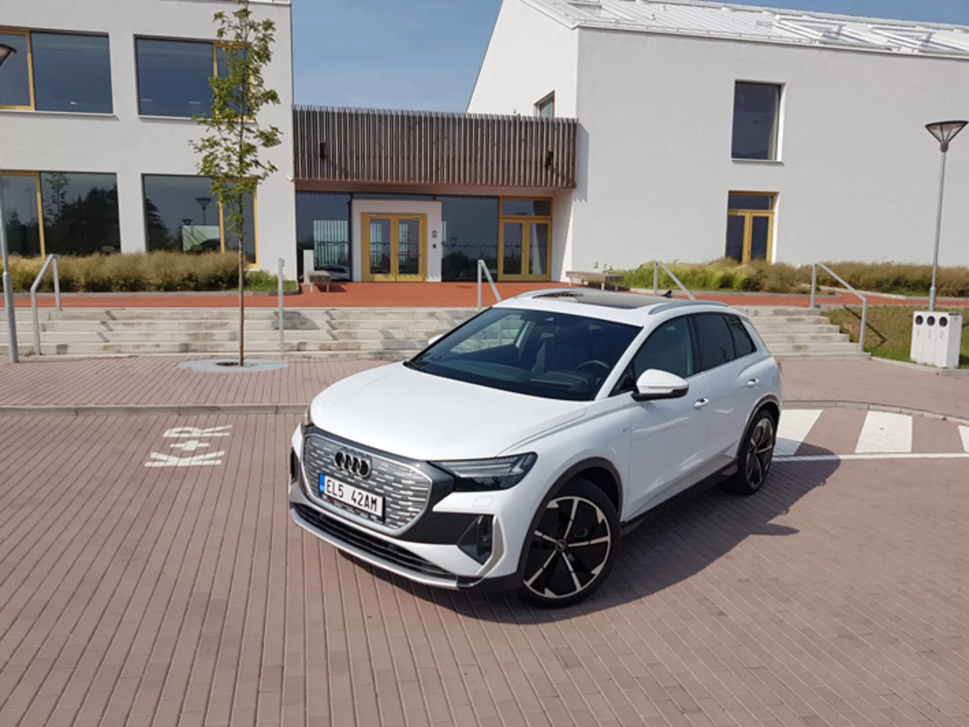 Bílý Audi Q4 e-tron 2021 s moderními LED světlomety, šestiúhelníkovou přední mřížkou a velkými slitinovými koly zaparkovaný na dlažbě s cihlovým vzorem s městskou architekturou a nabíjecí stanicí na pozadí pod jasnou polední oblohou.