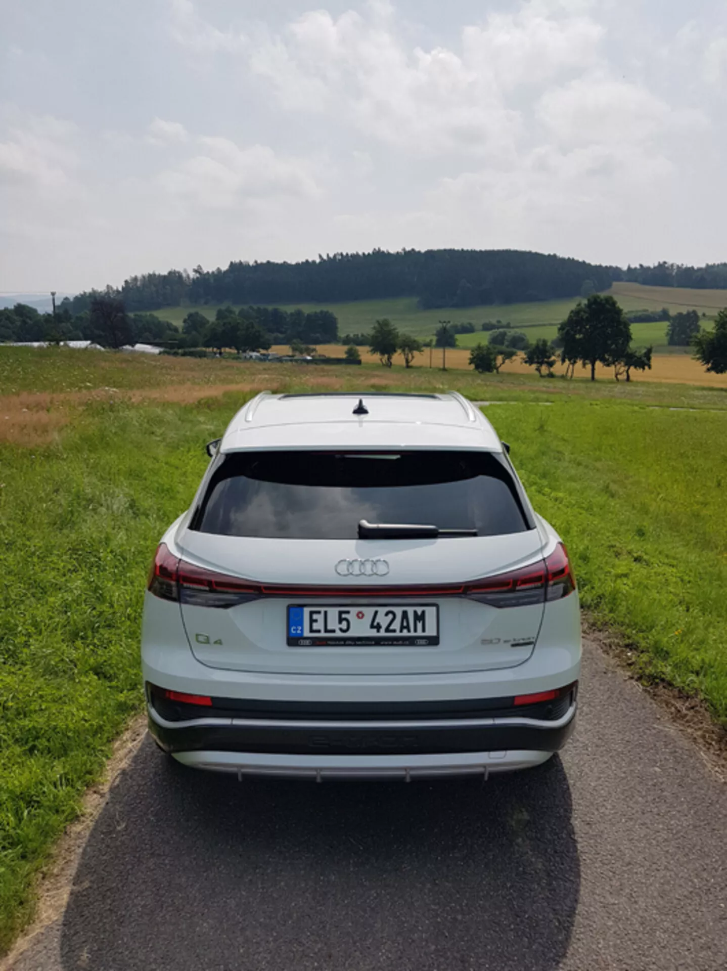 Zadní pohled na stříbrný Audi Q4 e-tron 2021 na úzké zpevněné cestě uprostřed pastorální krajiny s poli a stromy pod částečně zataženou oblohou, zdůrazňující jeho elegantní design, světelný pruh brzdových světel, střešní spoiler, senzory pro parkování vzadu a dvoubarevná slitinová kola.