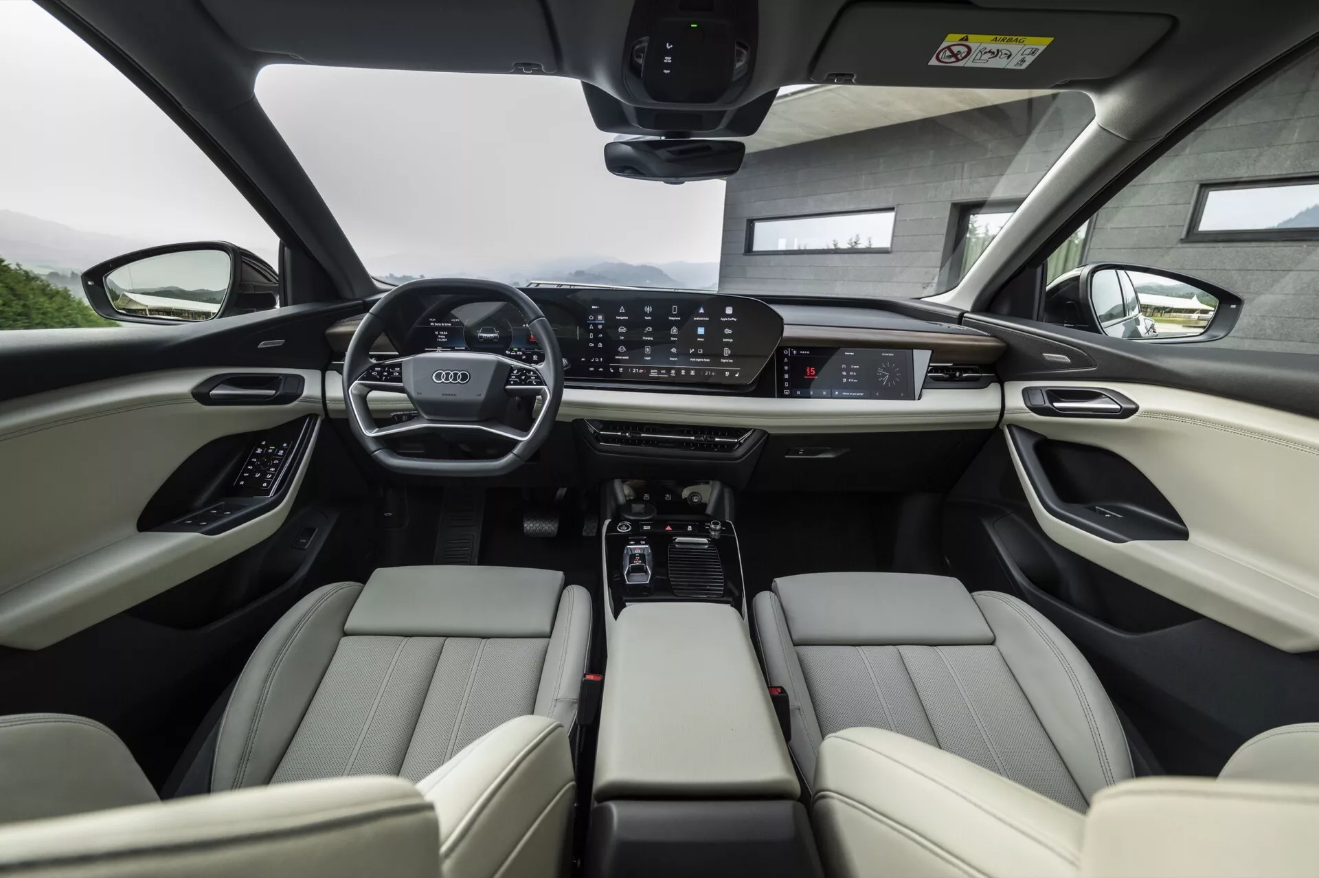 Pohled z interiéru Audi Q6 e-tron 2024 z perspektivy zadního sedadla zaměřený na přední řadu. Světle kožená přední sedadla jsou vybavena nastavitelnými opěrkami hlavy a elektronickými ovladači. Palubní deska obsahuje digitální přístrojový panel a velkou centrální dotykovou obrazovku pro infotainment, obklopenou ovládacími tlačítky pro klimatizaci a jízdní režimy. Středová konzola obsahuje držáky na nápoje, úložný prostor a automatickou převodovku. Volant je vybaven několika ovládacími prvky a panoramatická střecha umožňuje dostatek přirozeného světla. Obrázek zdůrazňuje pokročilé technologie a luxusní prvky automobilu.