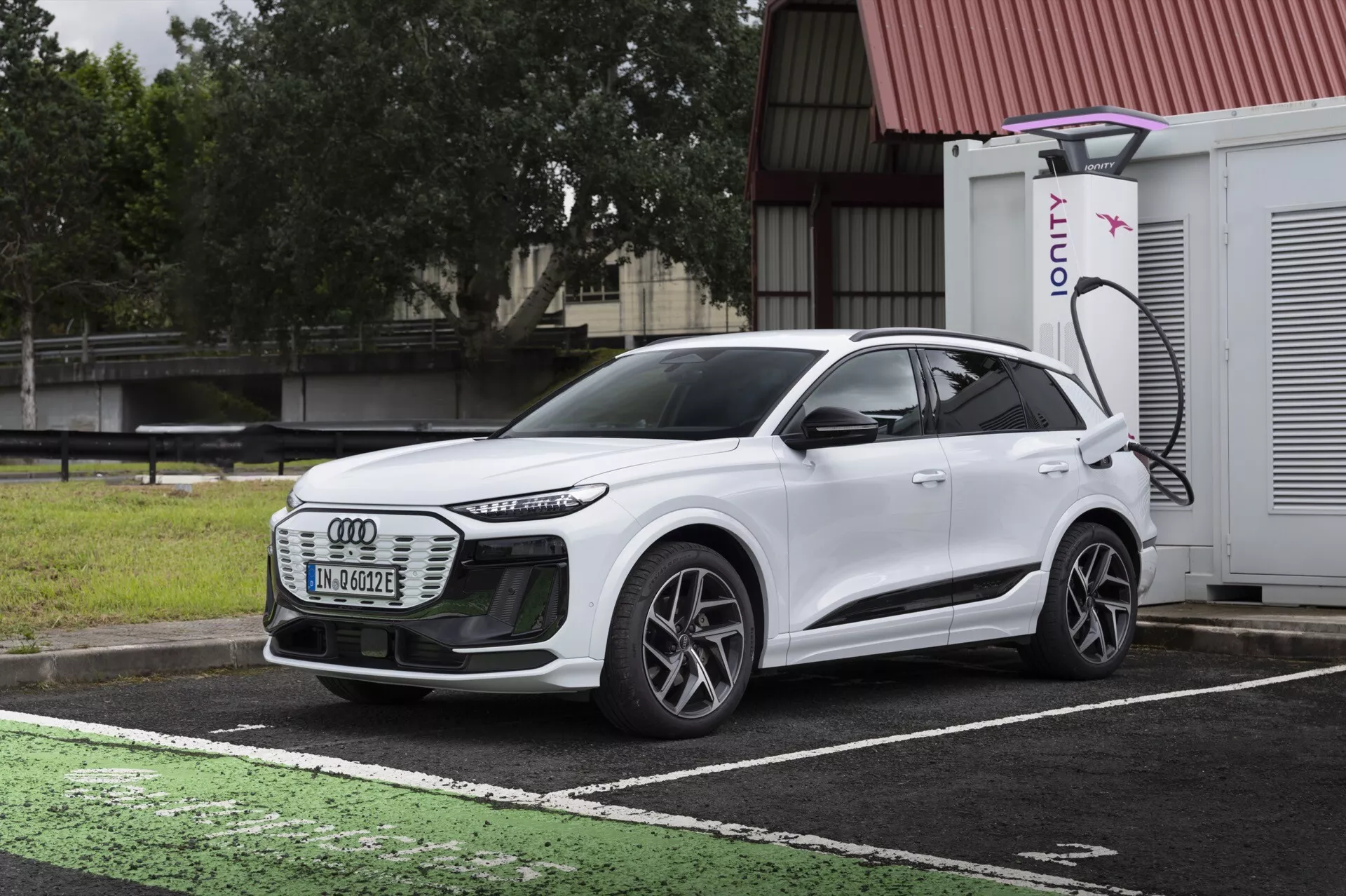Tříčtvrteční pohled zepředu na bílý elektrický SUV Audi Q6 e-tron 2024, zvýrazňující přední mřížku, LED světlomety, aerodynamické linie, vícepaprsková kola a boční profil. Vozidlo je zaparkováno na vyhrazeném místě pro nabíjení elektrických vozidel s viditelným nabíjecím portem a nabíječkou IONITY v pozadí. Obrázek kontrastuje design auta s tmavou, zataženou oblohou a mokrým chodníkem, ilustrující moderní technologii elektrických vozidel a pokročilé osvětlení.