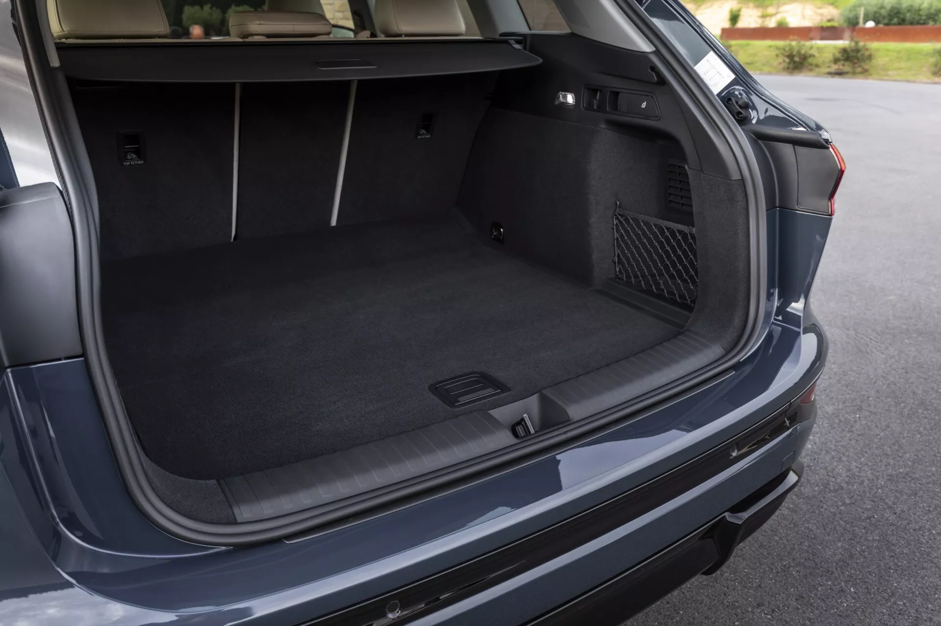Obrázek zobrazuje otevřený kufr moderního vozu Audi Q6 e-tron 2024, zachycený z úhlu mírně nad a napravo od zadní části vozu. Tento úhel poskytuje jasný pohled na prostorný, kobercem pokrytý zavazadlový prostor, zdůrazňující jeho hloubku a šířku. Významné prvky zahrnují tvarovanou přihrádku vlevo na drobné předměty nebo nouzové vybavení a síťovou kapsu vpravo. Na víku kufru je viditelné možné tlačítko pro automatické zavírání. Zadní nárazník a částečný pohled na pravé zadní světlo naznačují design exteriéru vozidla.