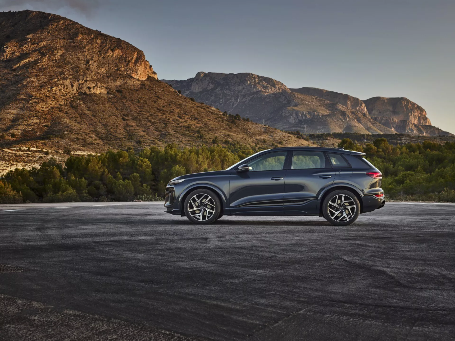 Moderní SUV, zřejmě Audi Q6 e-tron 2024, zaparkované venku s horským pozadím, zachycené z tříčtvrtečního zadního pohledu z vyvýšeného úhlu. Tmavá metalická barva karoserie kontrastuje s přírodním prostředím, čímž vynikají elegantní linie a aerodynamické kontury. Viditelné detaily zahrnují LED zadní světla, velká litá kola s multi-paprskovým designem, dvojitý výfukový systém, tónovaná okna, panoramatické střešní okno a ochranné lišty podél spodních okrajů. Zadní okno je mírně skloněné s zadním spoilerem a velkými postranními zrcátky s integrovanými směrovými světly, což zvyšuje sportovní a luxusní vzhled vozidla.
