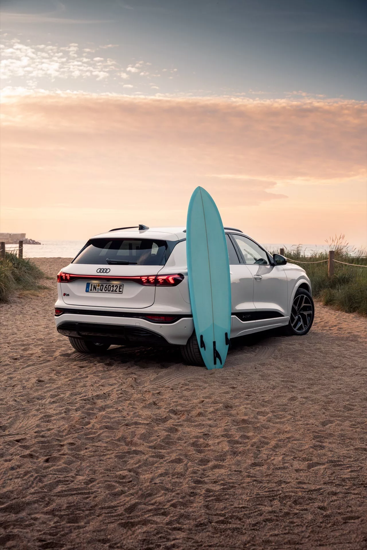 Obrázek zobrazuje bílé SUV Audi Q6 e-tron 2024 zaparkované na písčitém pobřeží s surfovacím prknem. Fotografie pořízená zezadu v tříčtvrtečním úhlu ukazuje plynulé linie vozu, LED zadní světla, parkovací senzory, kameru vzadu a vícepaprsková slitinová kola. Odraz zapadajícího slunce zvýrazňuje pokročilou kvalitu laku, zatímco surfovací prkno opřené o vůz naznačuje prvek dobrodružného životního stylu.