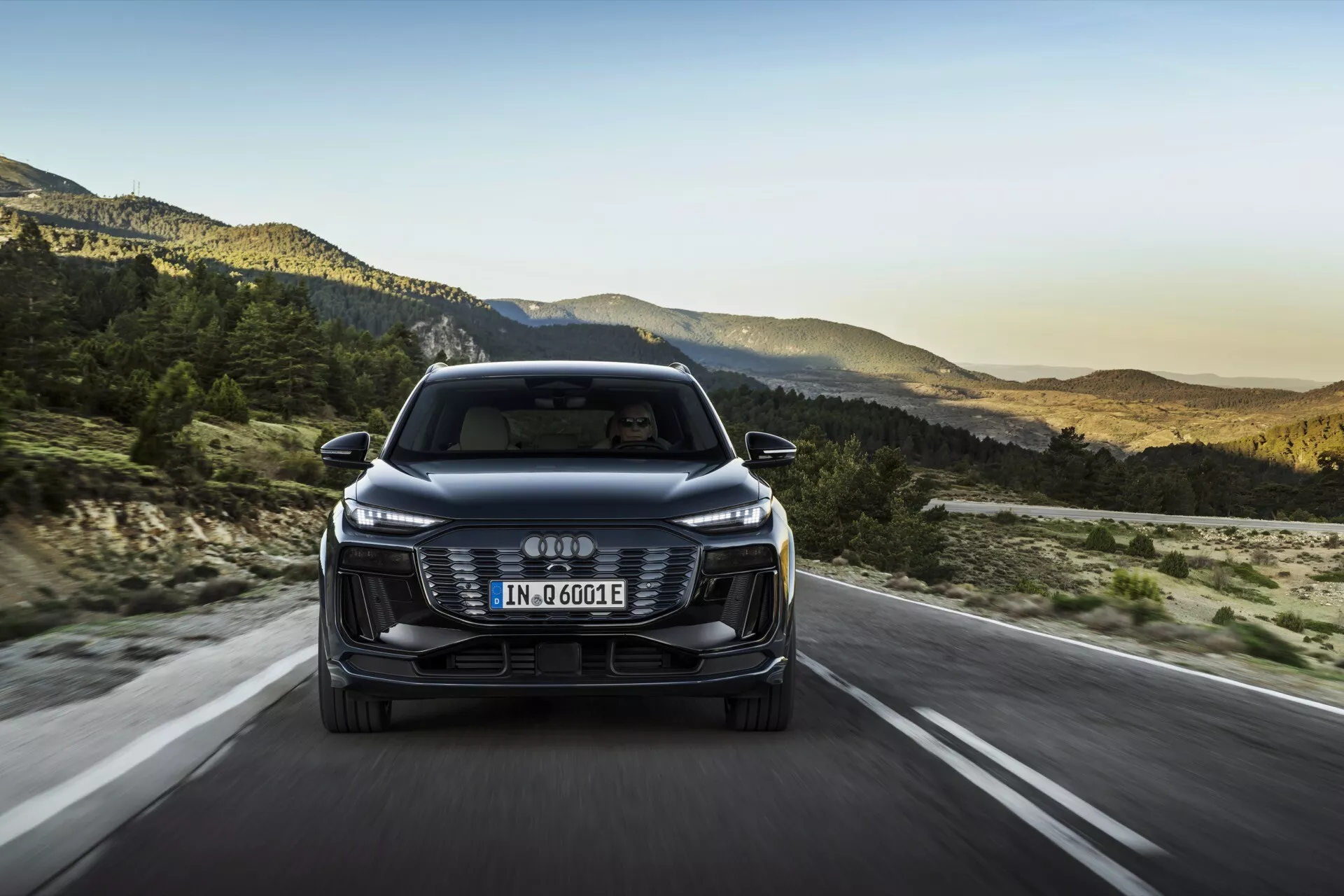Čelní pohled na Audi Q6 e-tron 2024, který ukazuje přední mřížku, světlomety a nárazník se čtyřmi vzájemně propojenými kruhy jako logem. Evropská registrační značka je umístěna na mřížce a označení 'e-tron' naznačuje jeho elektrickou/hybridní povahu. Auto jede po otevřené silnici s horskou krajinou v pozadí, což naznačuje nastavení pozdního odpoledne. Funkce zahrnují elegantní LED světlomety, estetickou mřížku, senzory/kamery asistence řidiče, aerodynamická zrcátka a efektivní design.