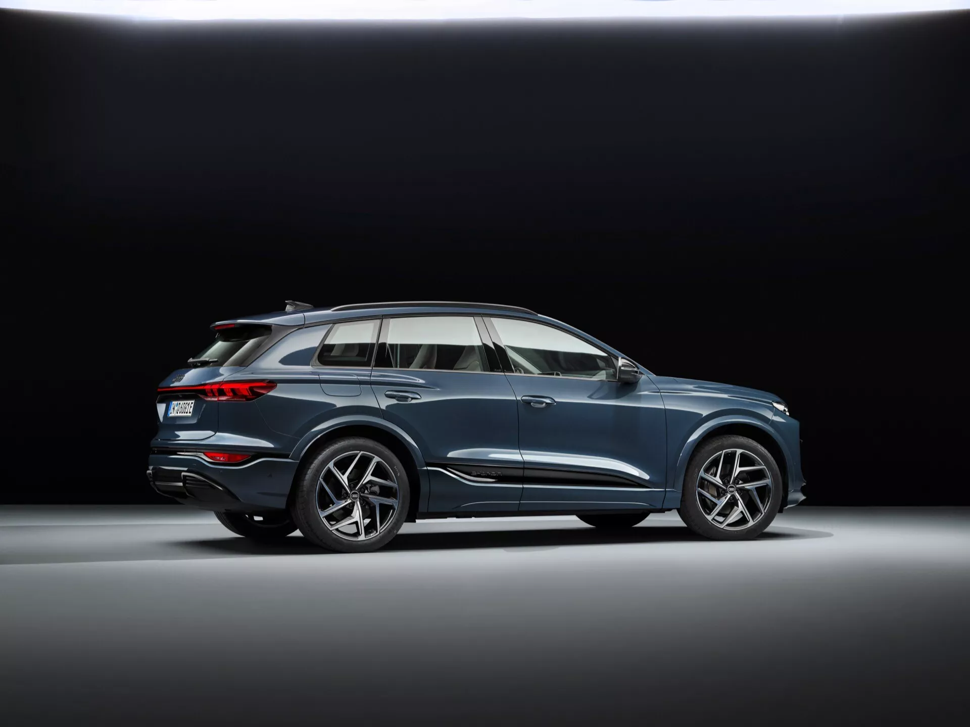 Nízký profilový záběr Audi Q6 e-tron 2024 ve studiu s tmavým pozadím, vyznačující se sportovními vícepaprskovými litými koly, plynulým designem karoserie s charakterovou linií, aerodynamickými bočními zrcátky s integrovanými směrovkami, chromovanými okenními rámečky, panoramatickým střešním oknem, střešními ližinami, střední jízdní výškou a zapuštěným dvojitým výfukovým systémem.