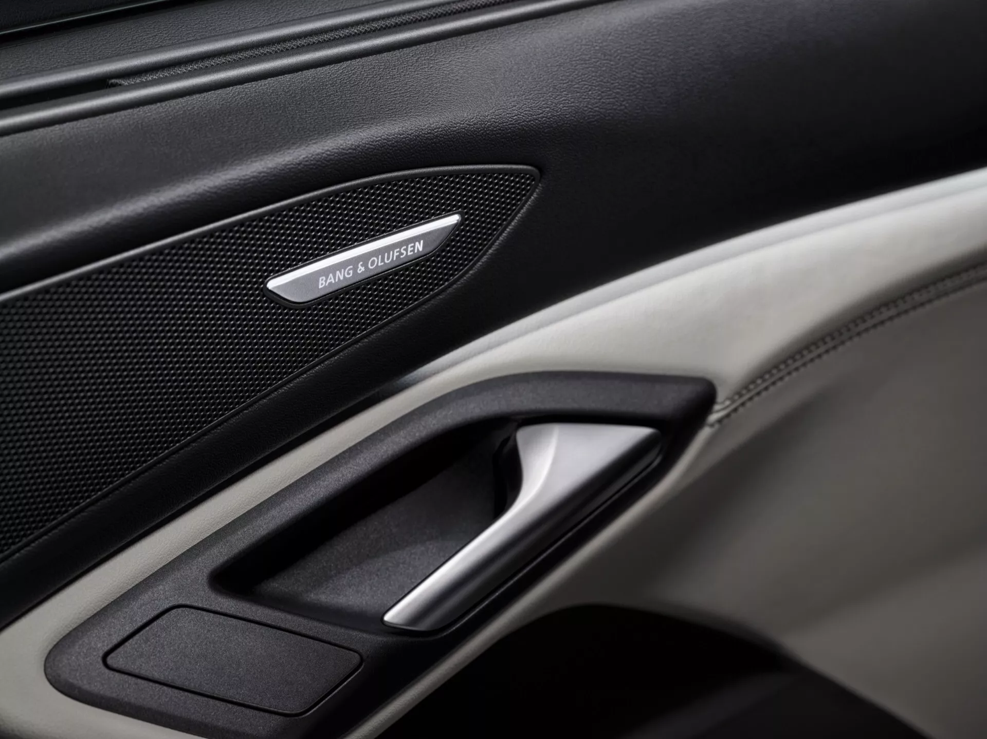 Detailní záběr na interiérový dveřní panel Audi Q6 e-tron 2024 GF, zdůrazňující prémiové prvky jako je mřížka reproduktoru Bang & Olufsen, pochromované dveřní madlo a kombinace kůže a textur z uhlíkových vláken. Dobře osvětlený obrázek se zaměřuje na detailní prvky panelu, prezentující luxusní a technologické prvky vozidla.