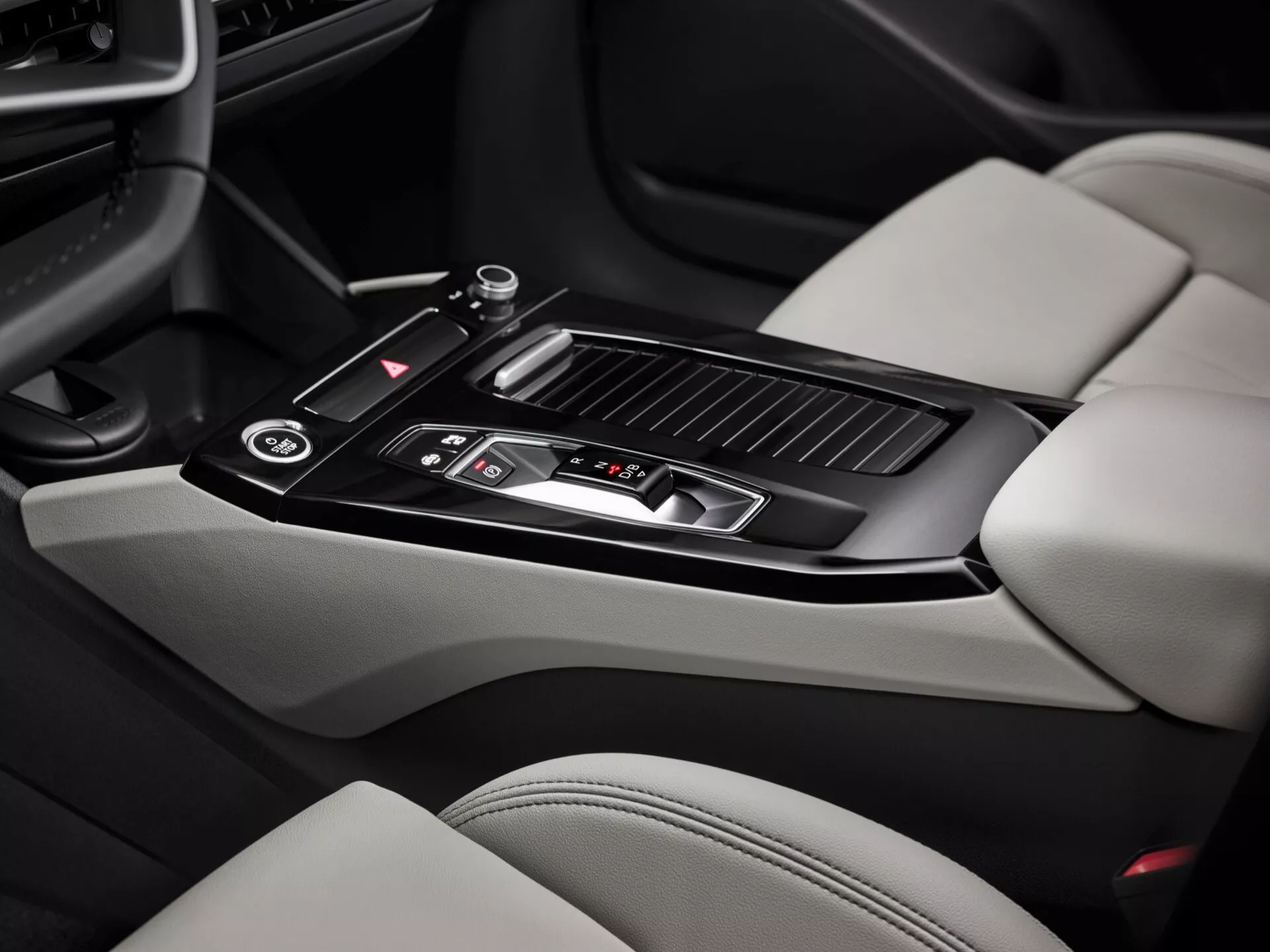Snímek interiéru Audi Q6 e-Tron 2024, zaměřený na oblast středové konzole. Obraz, pořízený z perspektivy spolujezdce, zdůrazňuje lesklou černou řadicí páku s kovovými detaily, několik ovládacích tlačítek, malý úložný prostor s posuvným krytem, dva držáky na nápoje, koženou opěrku paže, větrací otvory klimatizace a části světle zbarvených kožených sedadel s kontrastními tmavšími sekcemi. Funkce středové konzole ukazují moderní design a luxusní prvky.