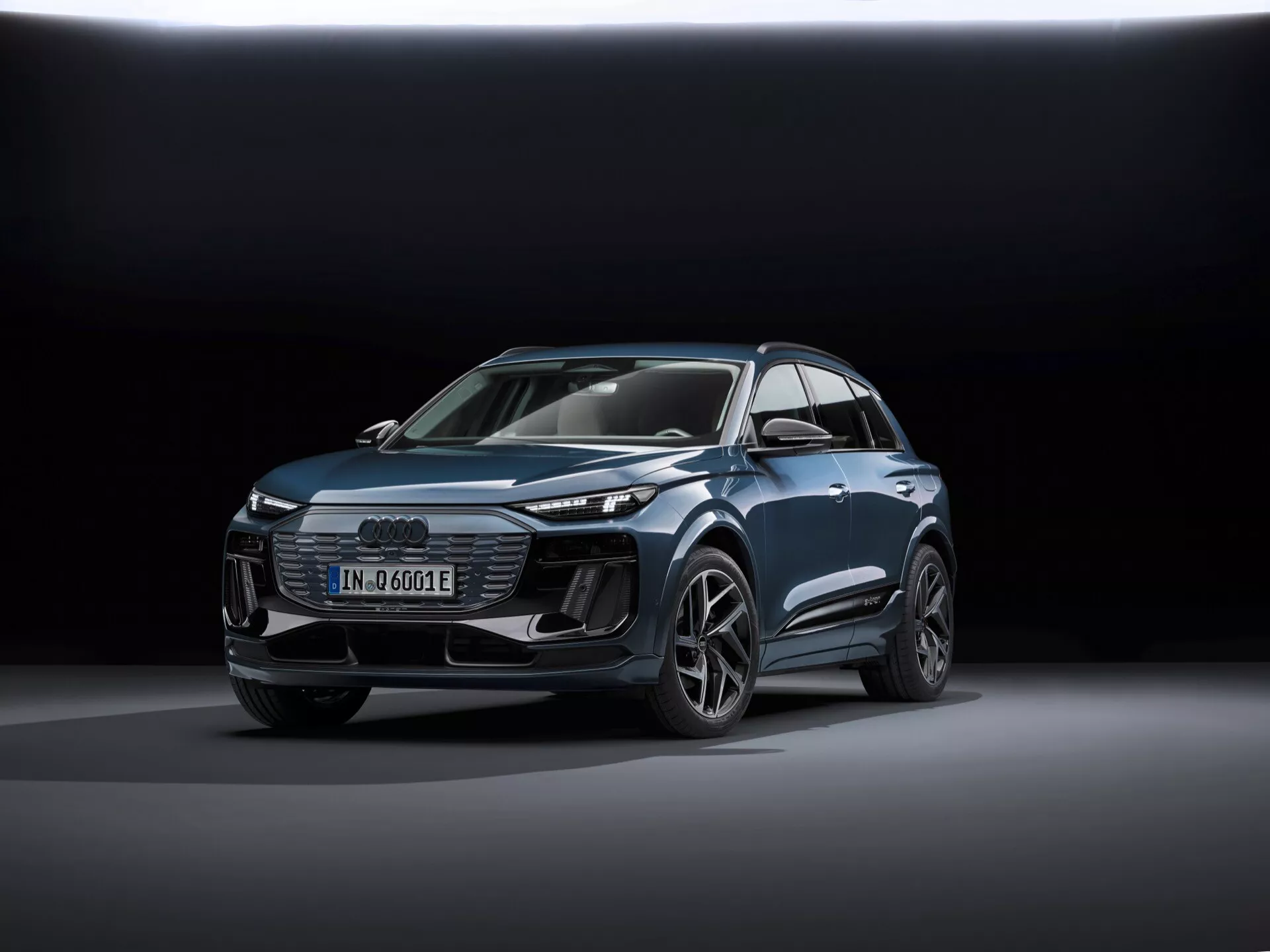 Obrázek zobrazuje moderní SUV Audi Q6-e-tron 2024 ve tříčtvrtinovém předním pohledu, zdůrazňujícím jak čelní, tak boční profil. Klíčovými identifikátory jsou čtyři kruhy znaku Audi na mřížce a odznak 'e-tron', indikující elektrický pohon. Snímek je pořízen z nízkého úhlu, což zvyšuje siluetu vozidla a zobrazuje jeho designové linie, svalnaté oblouky kol a aerodynamické prvky. Auto je umístěno ve středu rámce, stojí na hladkém reflexním povrchu s měkkým, rozptýleným osvětlením a temným přechodovým pozadím, které zdůrazňuje design vozidla. Mezi viditelnými technologickými prvky jsou LED světlomety, denní svícení (DRL), digitální displej a složitě navržené slitinné disky. Celkové lesklé provedení naznačuje propagační fotografii, která zdůrazňuje technologii, design a luxusní vzhled vozidla.