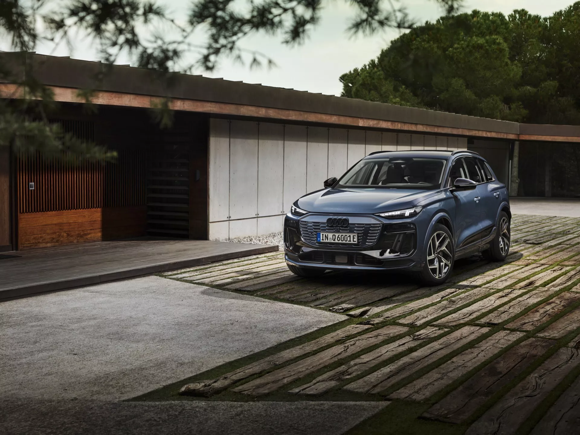 2024 Audi Q6 E-Tron tříčtvrteční přední pohled SUV s elegantními LED světlomety, výraznou mřížkou a velkým emblémem zaparkovaným na vzorované dlažbě. Luxusní budova a stromy v pozadí zdůrazňují luxus a exkluzivitu.