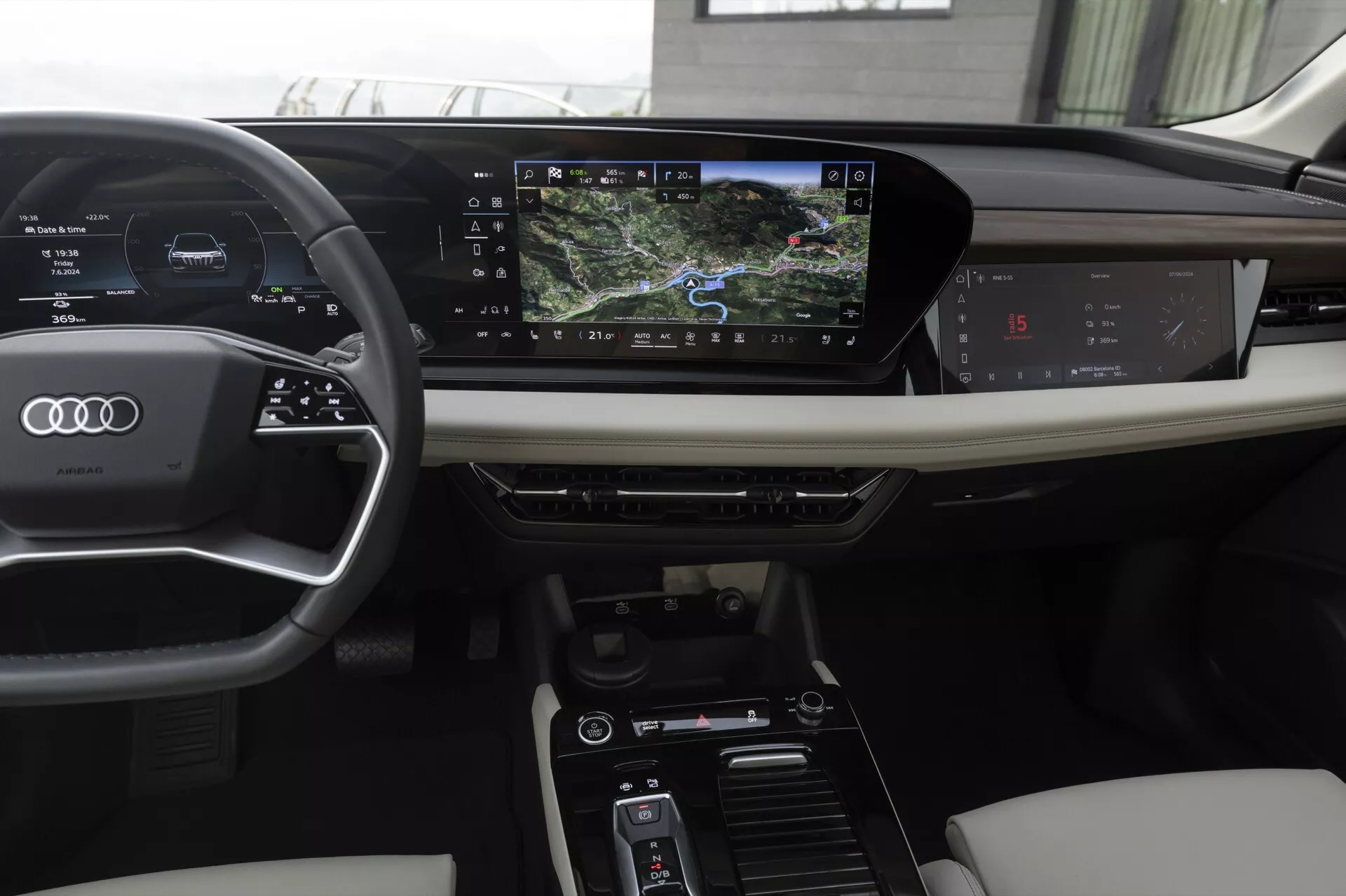 Interiér moderního Audi Q6 e-tron 2024, zobrazený z pohledu řidiče. Vybavení zahrnuje potažený volant s třemi rameny, ovládací prvky, digitální přístrojový panel zobrazující rychlost a navigaci, a horizontálně orientovaný dotykový infotainment systém. Středová konzole obsahuje ovládací prvky klimatizace a jízdních režimů. Luxusní materiály jako kůže a dřevěné obložení vylepšují estetiku.