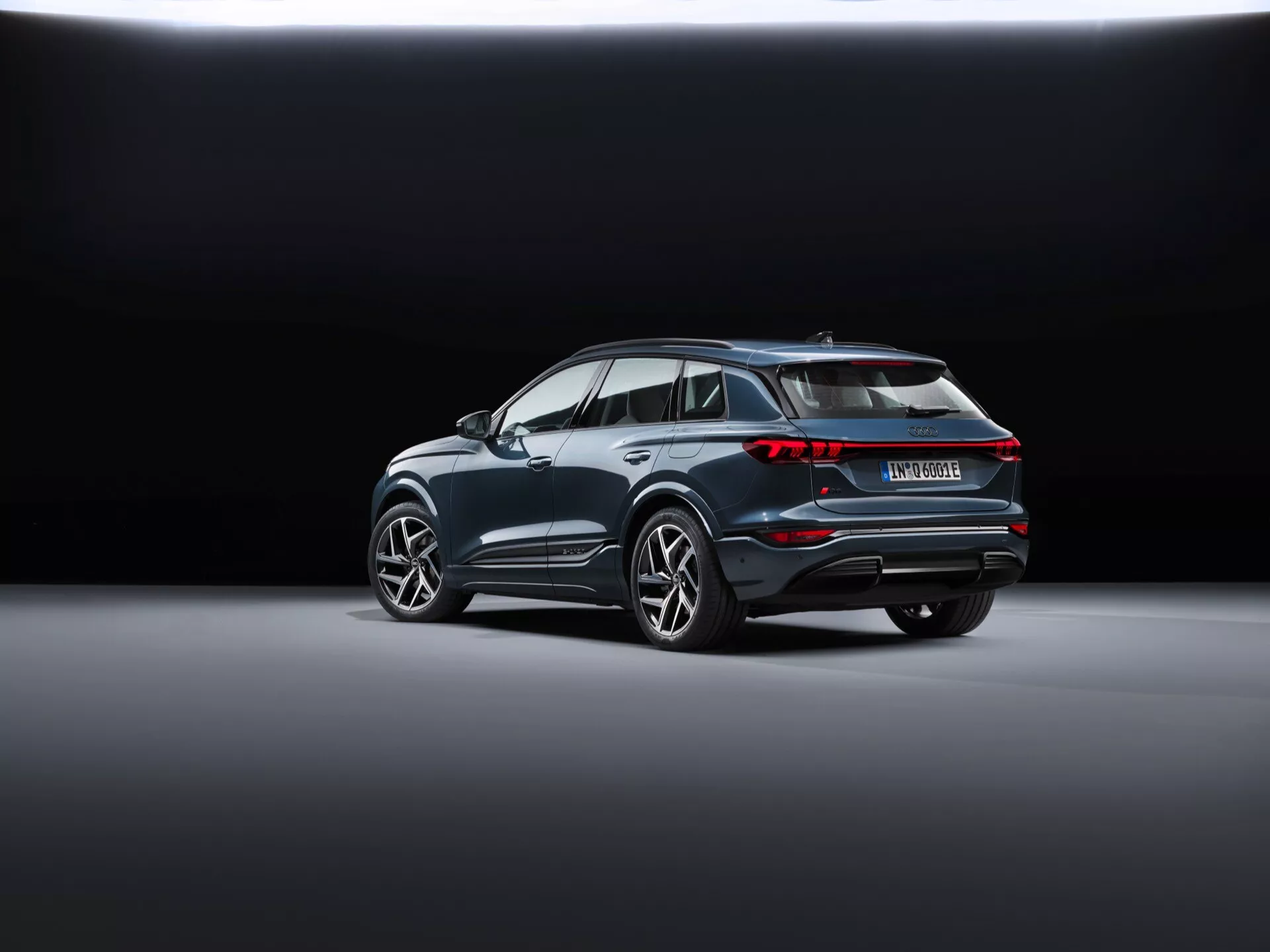 Zadní tříčtvrteční pohled na Audi Q6 e-tron 2024 (gf) ukazuje moderní SUV diagonálně umístěné na hladké, reflexní podlaze showroomu. Pozadí přechází z tmavé do světlé, čímž zdůrazňuje vozidlo. Viditelné prvky zahrnují propojené zadní světla, výrazný střešní spoiler, výrazné oblouky kol s velkými litými koly, chromovanou lištu podél oken, zadní nárazník s reflektory a senzory a dvojitý výfukový systém naznačující výkon, což může naznačovat, že se jedná o hybridní nebo elektrický model.