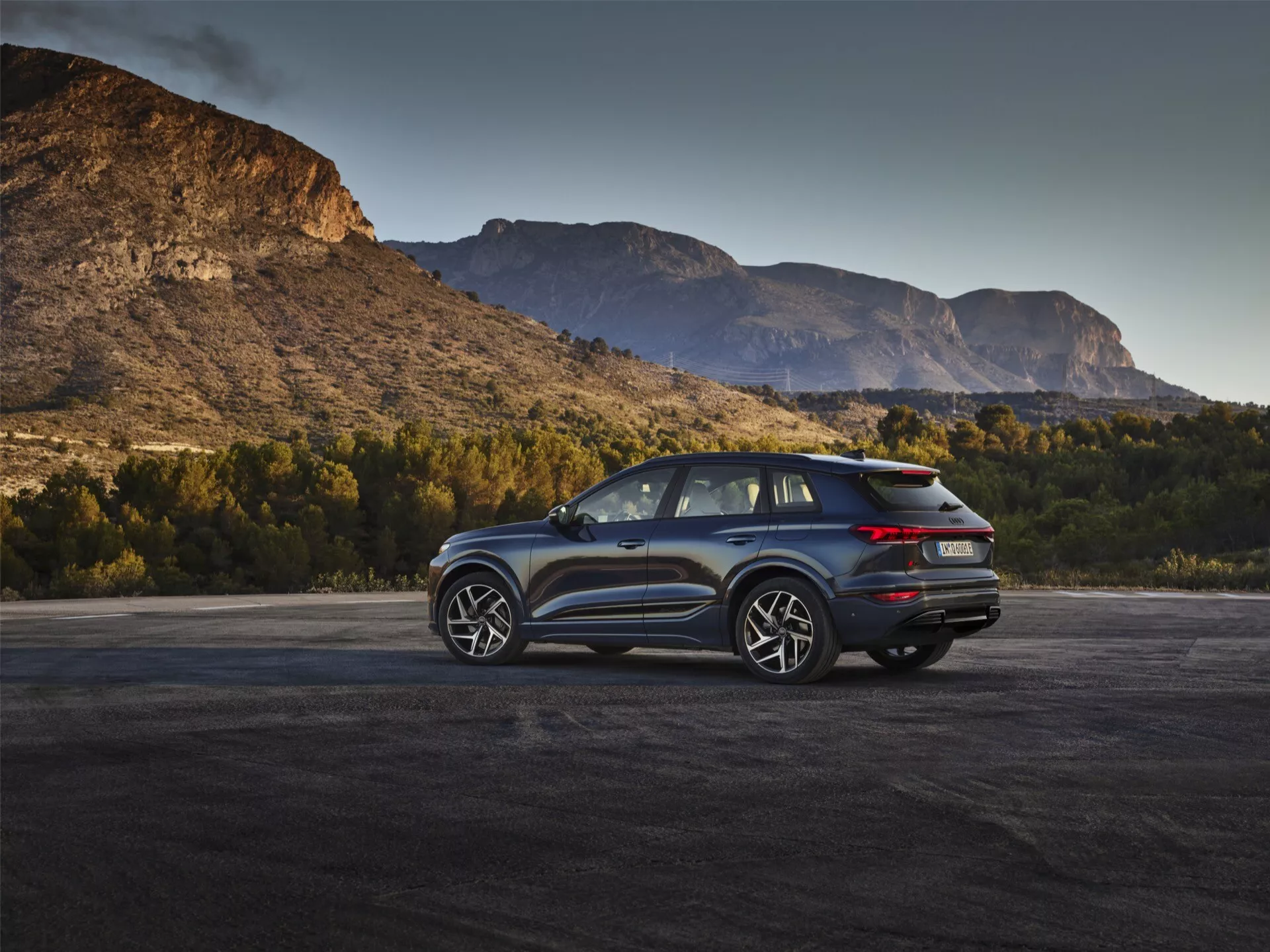 Obrázek zachycuje moderní SUV Audi Q6 e-Tron 2024 v přírodním prostředí s malebným horským pozadím za úsvitu nebo soumraku. Tříčtvrtečně zadní úhel zvýrazňuje výrazná LED zadní světla, čistý design zadního víka s možnou zadní kamerou a dvojitý výfukový systém. Boční profil ukazuje vícepaprsková litá kola, velká okna a skloněnou linii střechy, což naznačuje sportovní nebo luxusní výbavu s možnými aerodynamickými funkcemi. Zrcátka mohou integrovat technologii sledování slepých úhlů. Přirozené osvětlení zvyšuje přitažlivost vozidla v souvislosti s dobrodružstvím a schopnostmi.
