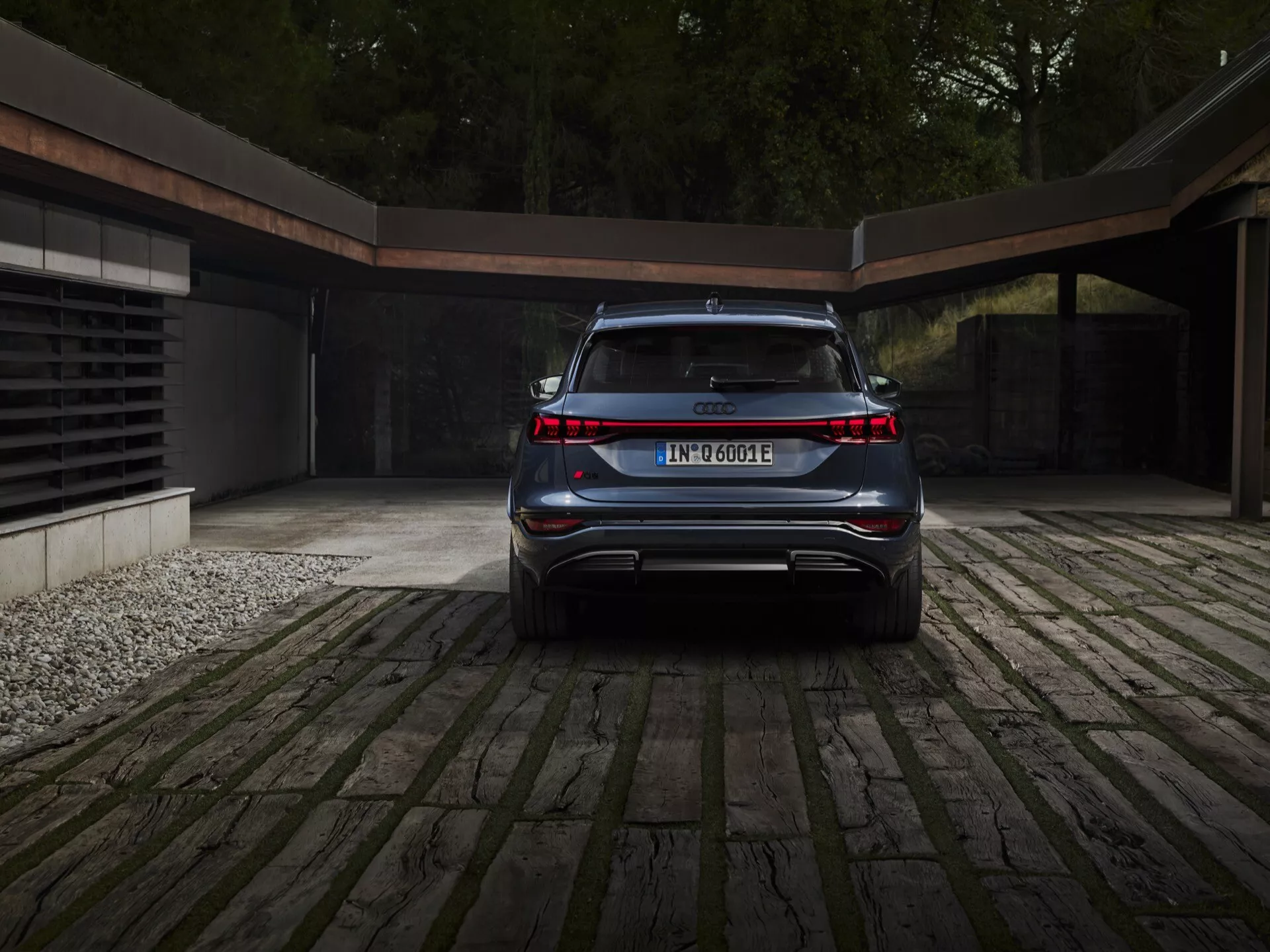 Pohled na zadní část vozu Audi Q6 e-tron 2024, zdůrazňující jeho čistý, elegantní a moderní design. Klíčové prvky, které jsou vidět, zahrnují spojitý světelný pás propojící obě zadní světla, označení modelu a elektrického pohonného ústrojí, integrovaný zadní nárazník se sportovním difuzorem, dopředu se svažující zadní okno naznačující SUV nebo crossover, skryté koncovky výfuku a středově umístěnou registrační značku. Vůz je zaparkován na dlážděném příjezdovém cestě s náznakem moderní garáže v pozadí, což naznačuje luxusní a ekologický životní styl.