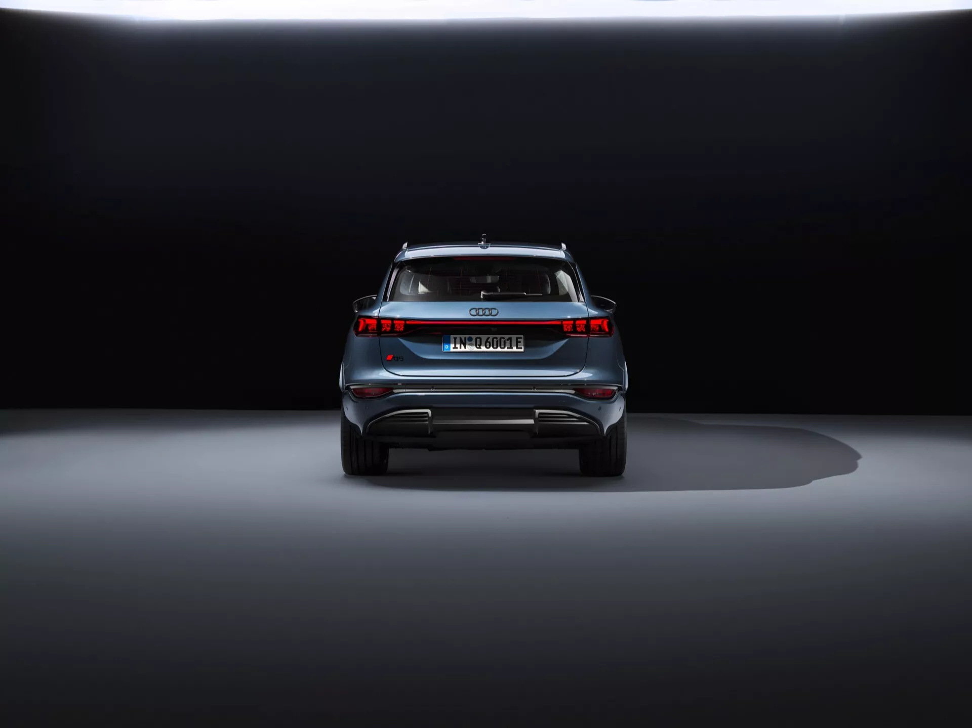 Zadní pohled na Audi Q6 e-tron 2024 na výstavě s dramatickým nasvícením. Snímek z úrovně země přibližuje moderní LED zadní světla, absence výfukových trubek, celoplošné osvětlení, evropskou poznávací značku, střešní spoiler a reflexní bezpečnostní prvky, což naznačuje, že se jedná o luxusní elektromobil nebo hybrid