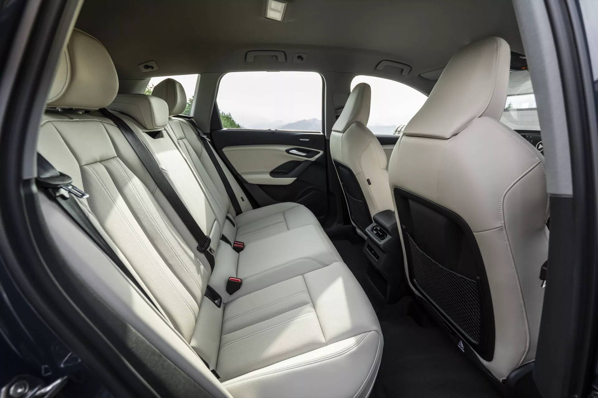 Interiér zadního prostoru pro cestující modelu Audi Q6 e-tron 2024, zaměřený na uspořádání zadních sedadel. Lavicová sedadla jsou potažena světle barevným koženým čalouněním s opěrkami hlavy, opěrkou a viditelnými bezpečnostními pásy. Na dveřním panelu je mřížka reproduktoru, klika dveří a pravděpodobně ovládání oken. Barevné schéma je dvoubarevné, se světlými a tmavými prvky. Mezi předními sedadly jsou větrací otvory klimatizace, což naznačuje klimatizaci pro zadní pasažéry. Moderní, luxusní a příjemný design pro cestující.