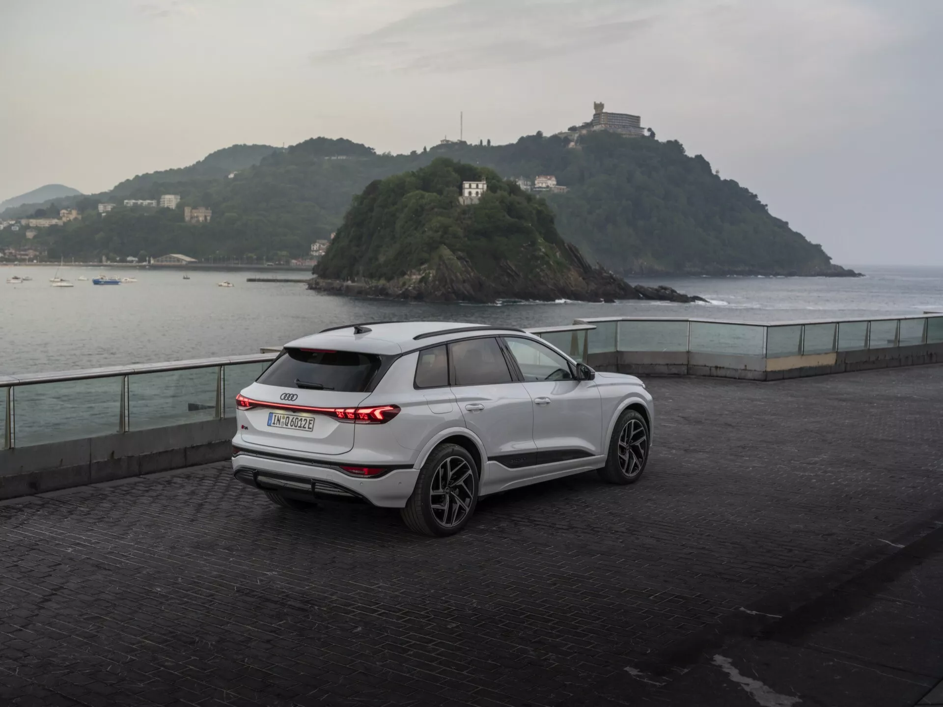 Tříčtvrteční zadní pohled na moderní bílý Audi Q6 e-tron 2024 SUV zaparkovaný na nábřeží při soumraku, ukazující jeho horizontální LED zadní světla, značku výrobce, stěrač zadního okna, pětipaprsková litá kola, integrované směrové ukazatele, střešní ližiny a tvarované linie karoserie proti poklidnému pozadí s malým ostrovem a budovou, zdůrazňujícím směs užitkovosti a výkonu.