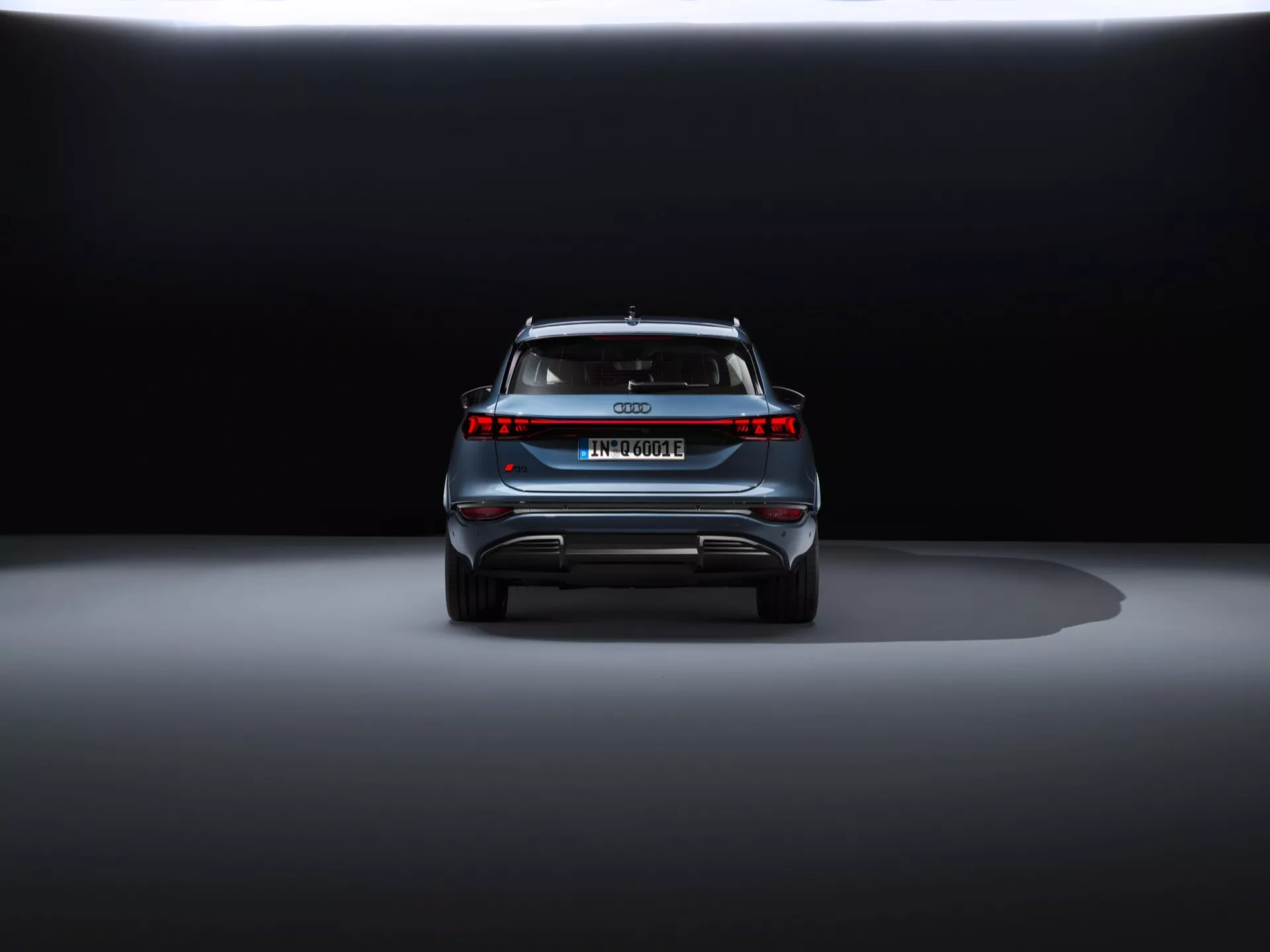 Zadní pohled na Audi Q6 e-tron 2024 na tmavém pozadí, předvádějící elegantní design, centrální znak, název modelu, horizontální světelnou lištu, zadní spoiler a difuzorovitý nárazník s reflexními prvky.