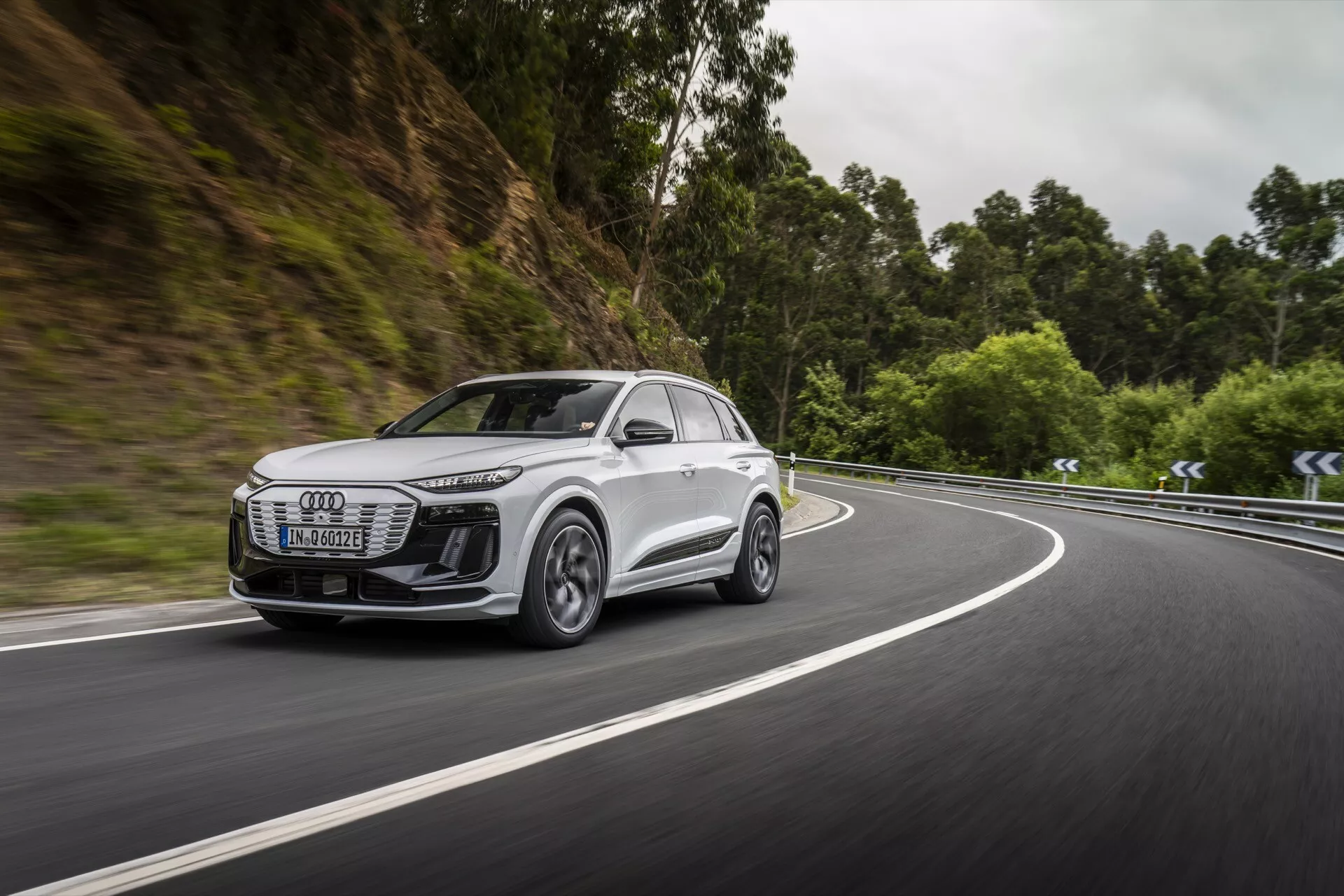 Obrázek zachycuje dynamický boční přední pohled na moderní bílé SUV, pravděpodobně model Audi Q6 e-tron 2024, jedoucí po silnici obklopené zalesněnou oblastí. Pozornost je zaměřena na přední masku, světlomety, boční profil, kola a aerodynamiku bočních zrcátek vozidla. Světlomety jsou elegantní s LED technologií, což naznačuje pokročilé osvětlení. Kola z lehkých slitin s vícepaprskovým designem naznačují vysokovýkonné schopnosti. Pohyb vozu na zakřivené silnici zdůrazňuje jeho jízdní vlastnosti a možnost pokročilých asistenčních systémů pro řízení. Rozmazané pozadí zdůrazňuje rychlost a efektivitu při navigaci v náročných situacích.