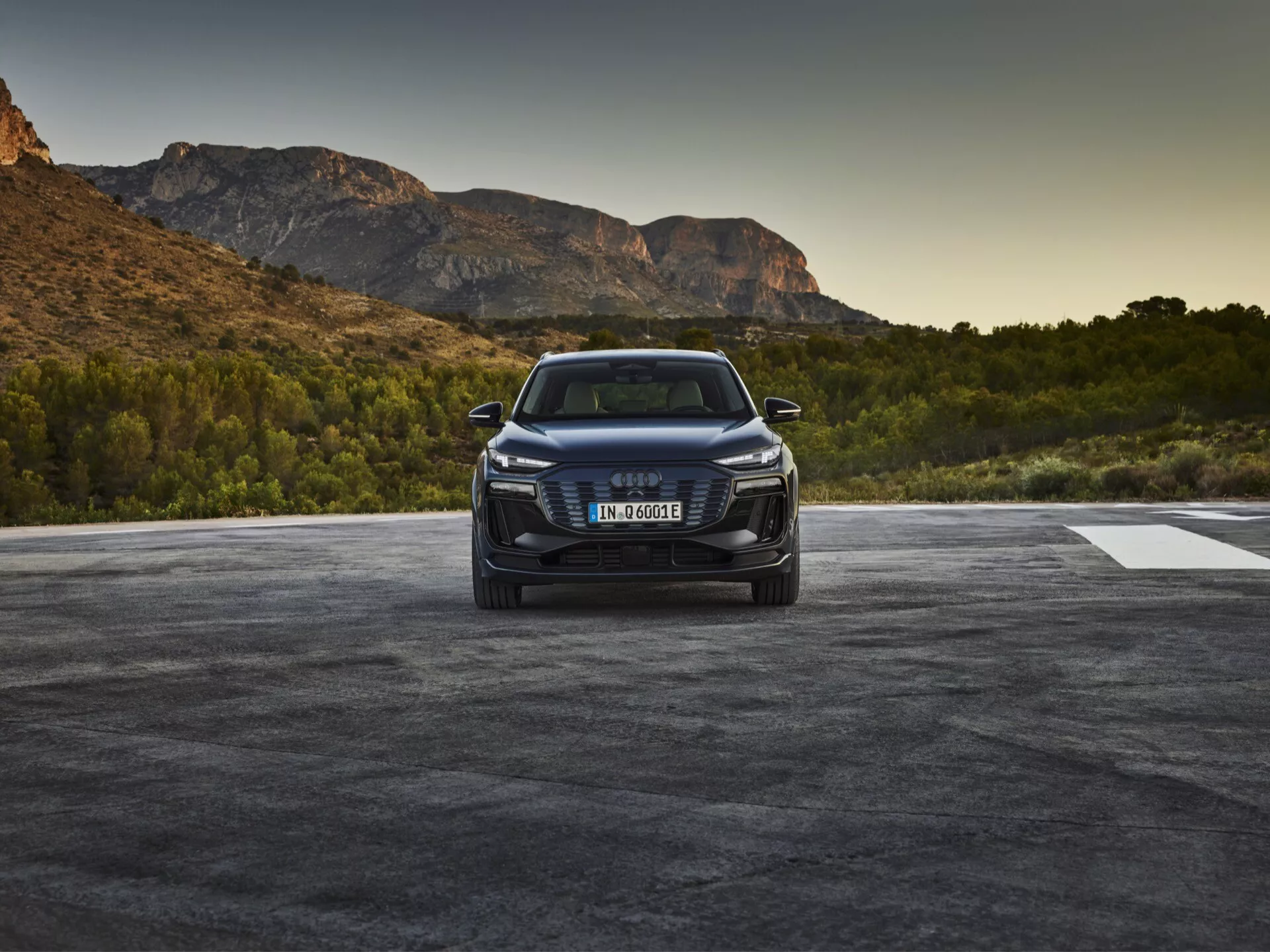Obrázek vozu Audi Q6 e-tron 2024 fotografovaného z předního tříčtvercového úhlu, který zobrazuje přední mřížku vozidla s emblémem značky, ostré LED nebo laserové světlomety a detailní přední nárazník s nasávacími otvory a možná senzory nebo kamerami naznačujícími asistenci při řízení. Kapota má obrysové linie a elegantní boční profil naznačuje aerodynamiku s vyvýšeným postojem SUV. Vůz je zasazen do venkovního prostředí s přírodní scenérií, které naznačuje východ nebo západ slunce.