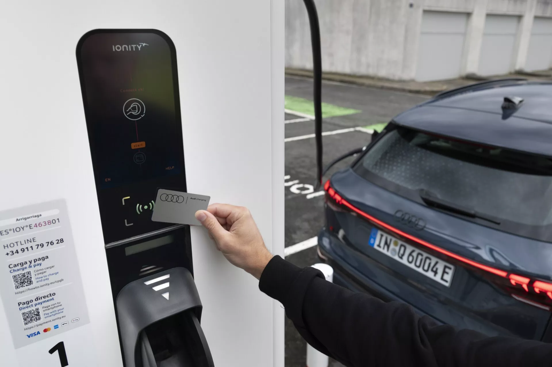 Obrázek osoby držící RFID kartu u čtečky nabíjecí stanice pro elektromobily IONITY. Zaměřeno na zadní část moderního, tmavě zbarveného automobilu s horizontálními LED světlomety, což naznačuje jeho elektrickou povahu. Auto, pravděpodobně EV, je zaparkováno vedle nabíječky, což naznačuje, že se buď nabíjí, nebo se připravuje na nabíjení.