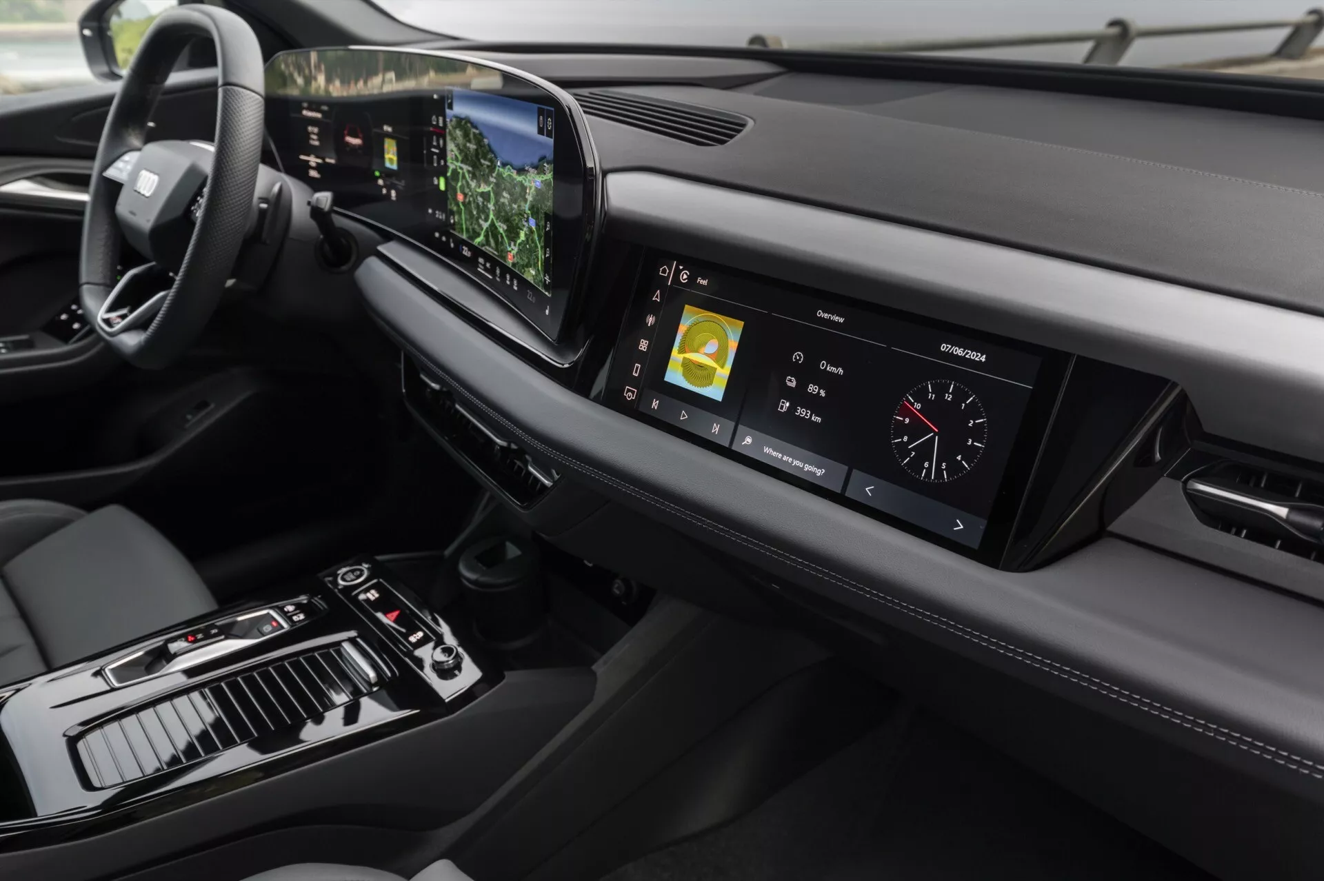 Pohled na interiér Audi Q6 e-tron 2024 zaměřený na palubní desku a centrální konzolu z mírně zvýšeného úhlu. Vlevo je viditelný volant, což naznačuje konfiguraci pro jízdu vlevo, zatímco centrální konzola obsahuje řadicí páku, různé ovládací prvky a velký dotykový displej. Digitální přístrojový klastr zobrazuje jízdní metriky. Stylová palubní deska se horizontálně rozprostírá s větracími otvory a dalším zobrazovacím prvkem vpravo. Pokročilá technologie je zřejmá z centrálního dotykového displeje zobrazujícího navigační a multimediální informace, projekčního displeje viditelného nad volantem a dotykových tlačítek pro ovládání klimatu. Design klade důraz na minimalismus a prémiové materiály, přičemž kožené šití a kovové akcenty přispívají k luxusnímu dojmu.