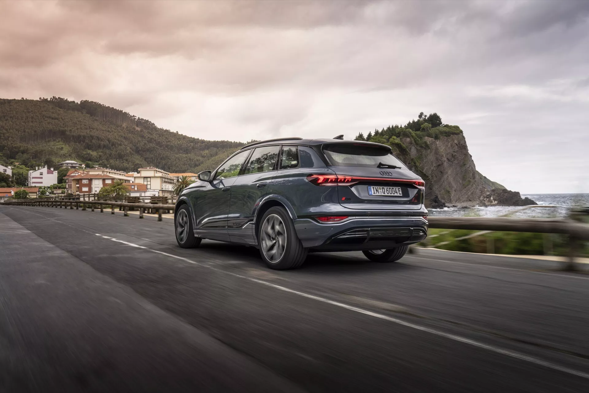 Audi Q6 e-tron 2024 z zadního tříčtvrtečního pohledu v pohybu na pobřežní silnici, předvádějící výrazná LED zadní světla, tmavě kovový lak, litá kola a praktický zadní stěrač. Rozmazané pozadí naznačuje pobřežní prostředí a odráží estetiku a technologický pokrok moderního SUV.