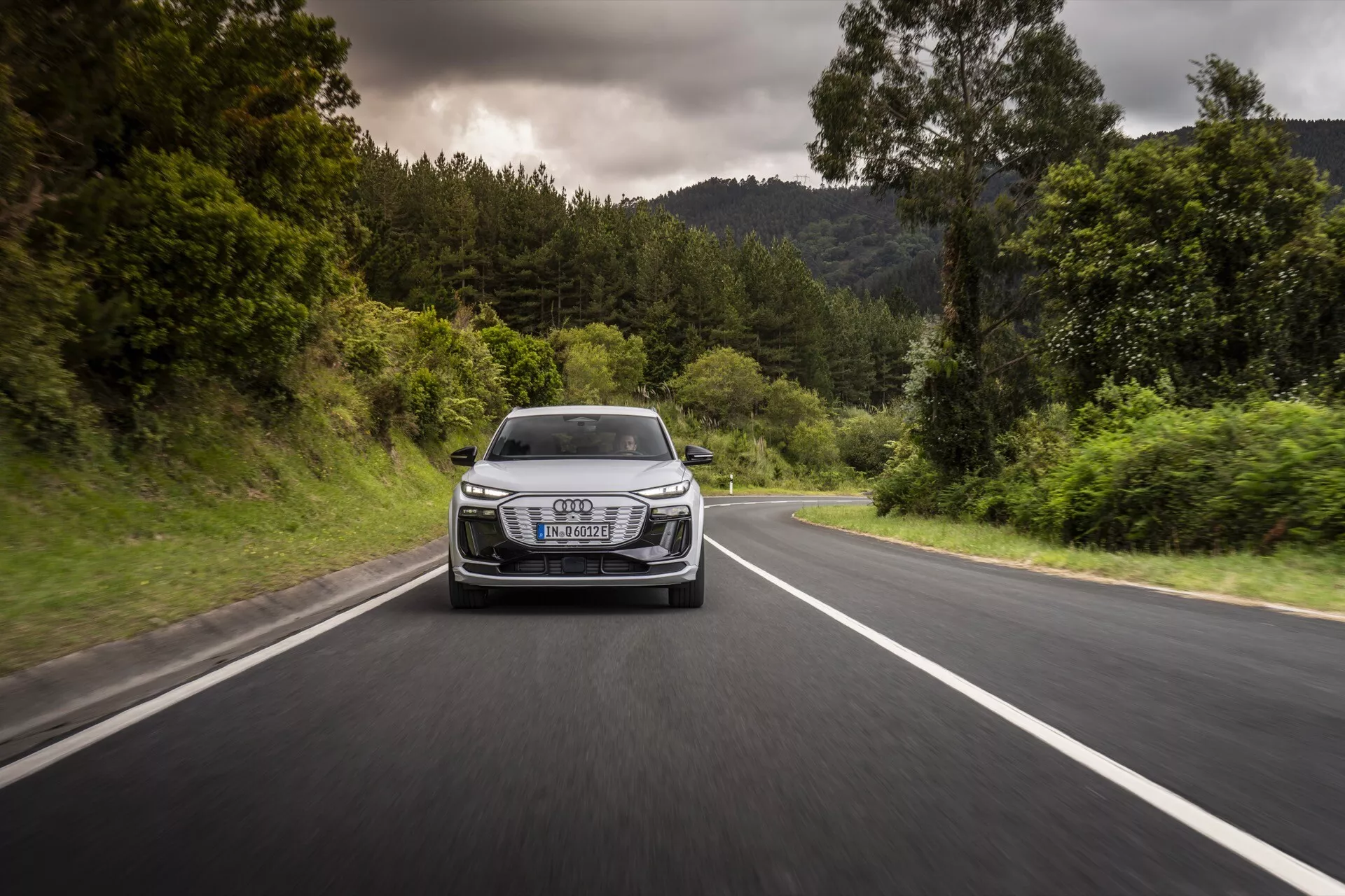Audi Q6-e-tron 2024 zachycený v pohybu z čelního úhlu na dvouproudé silnici lemované zelení. Fotografie ukazuje mřížku, světlomety, kapotu a nárazník vozidla, zdůrazňuje dynamický výkon a design. Vůz má charakteristické LED světlomety, výrazný luxusní znak na mřížce a aerodynamické linie, vše za oblačného počasí s suchými podmínkami na silnici.