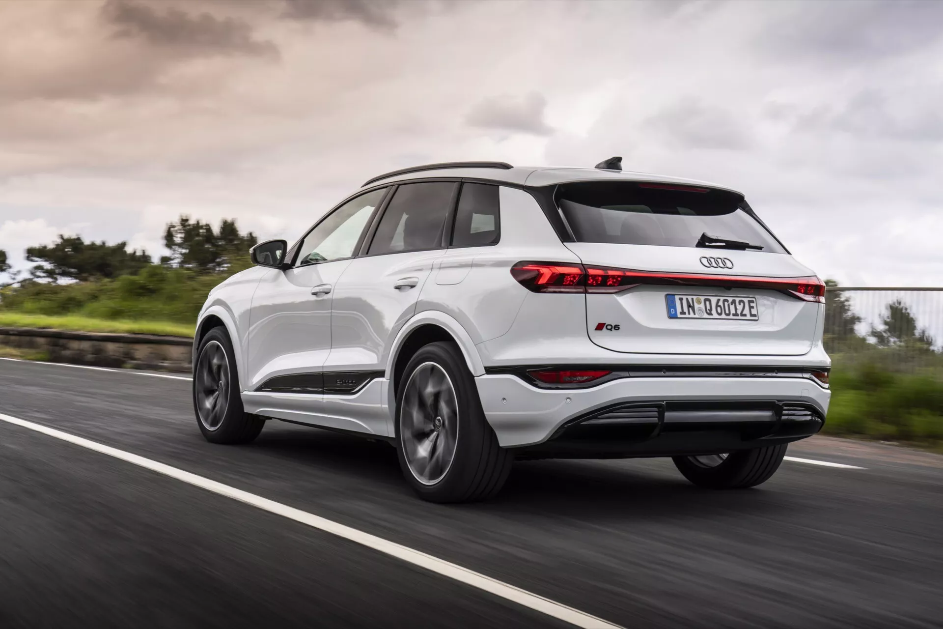 Audi Q4 e-tron 2024 jedoucí po asfaltové silnici zobrazený z pohledu zezadu z tři čtvrtiny, zachycující levou zadní stranu. Obrázek ukazuje úzké zadní světla, střešní spoiler, panoramatickou střechu, kola z lehké slitiny s vícepaprskovým designem, dvoubarevnou karoserii s kovovým lemováním a ikonický znak Audi. Rozmazaný pohyb kol a zelená kolem silnice s zataženou oblohou naznačují dynamické jízdní podmínky za různých počasí.