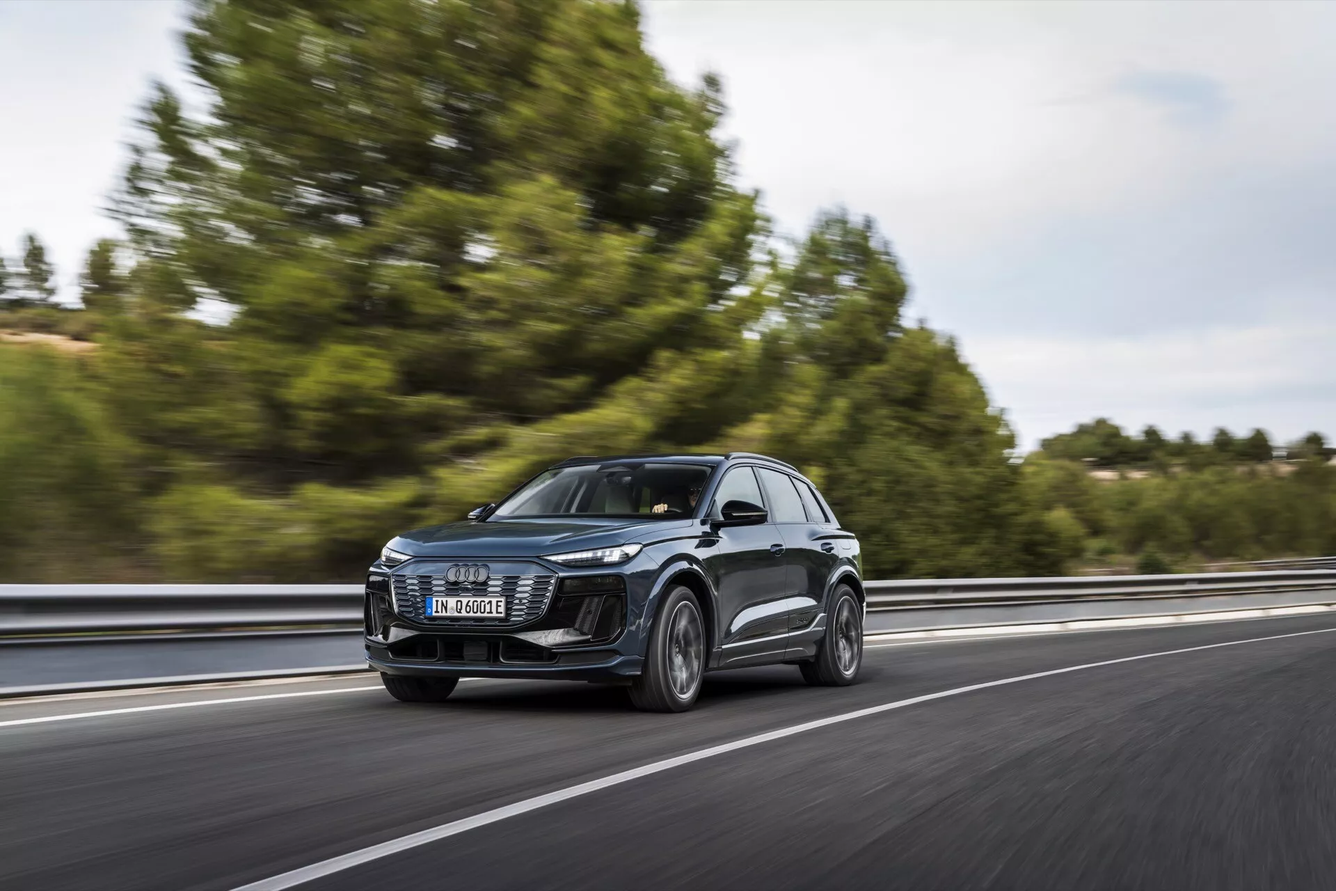 Audi Q6 e-tron 2024 SUV v pohybu na dálnici, zachycený z tříčtvrtečního předního pohledu. Obrázek ukazuje přední mřížku, LED světlomety, kapotu, čelní sklo a část bočního profilu, včetně dveří a bočních zrcátek. Vozidlo jede vpravo v pruhu, pohybuje se zleva doprava s rozmazaným pozadím naznačujícím rychlost. Stromy a svodidla jsou lehce viditelné. Jasný design předních světlometů naznačuje pokročilé osvětlovací systémy a velká mřížka naznačuje technologie chlazení nebo senzory pro ADAS. Boční zrcátka mohou obsahovat indikátory směru nebo systémy monitorování mrtvého úhlu. Kola poskytují vhled do kategorií výkonu nebo luxusu.