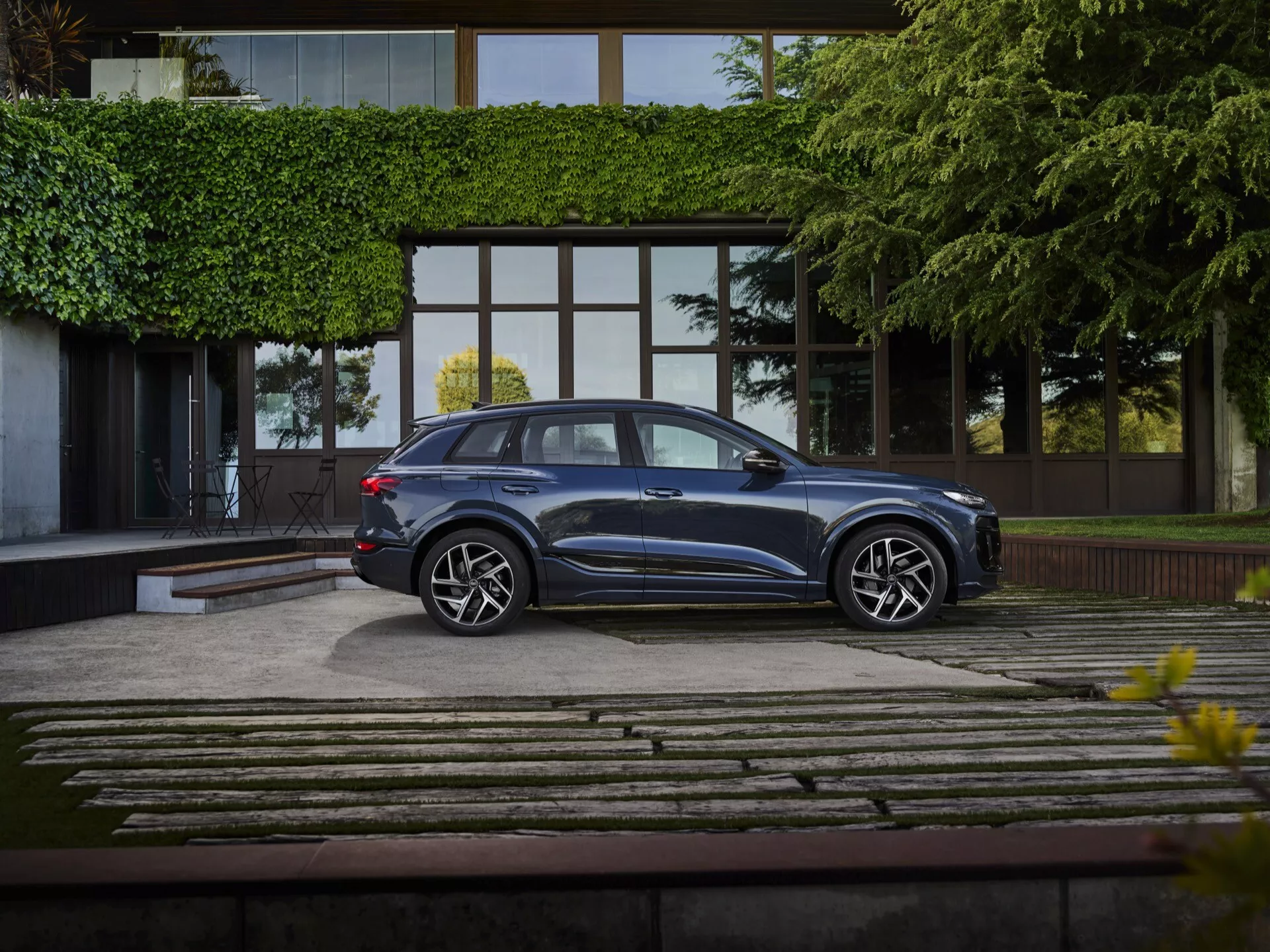 Na dobře upravené příjezdové cestě stojí Audi Q6 e-tron 2024, zachycené v kompletním profilovém pohledu z mírně zvýšeného úhlu. Hluboce kovově modré SUV se leskne elegantním designem, s aerodynamickými liniemi střechy, tónovanými okny a zapuštěnými klikami dveří. Vícepaprsková kola z lehké slitiny a nízkoprofilové pneumatiky zdůrazňují jeho špičkový výbavový stupeň. Na pozadí moderní luxusní budovy s velkými skleněnými okny a kamenami a trávníkovou příjezdovou cestou, je scéna jemně osvětlená, zvýrazňující prémiový status vozidla.