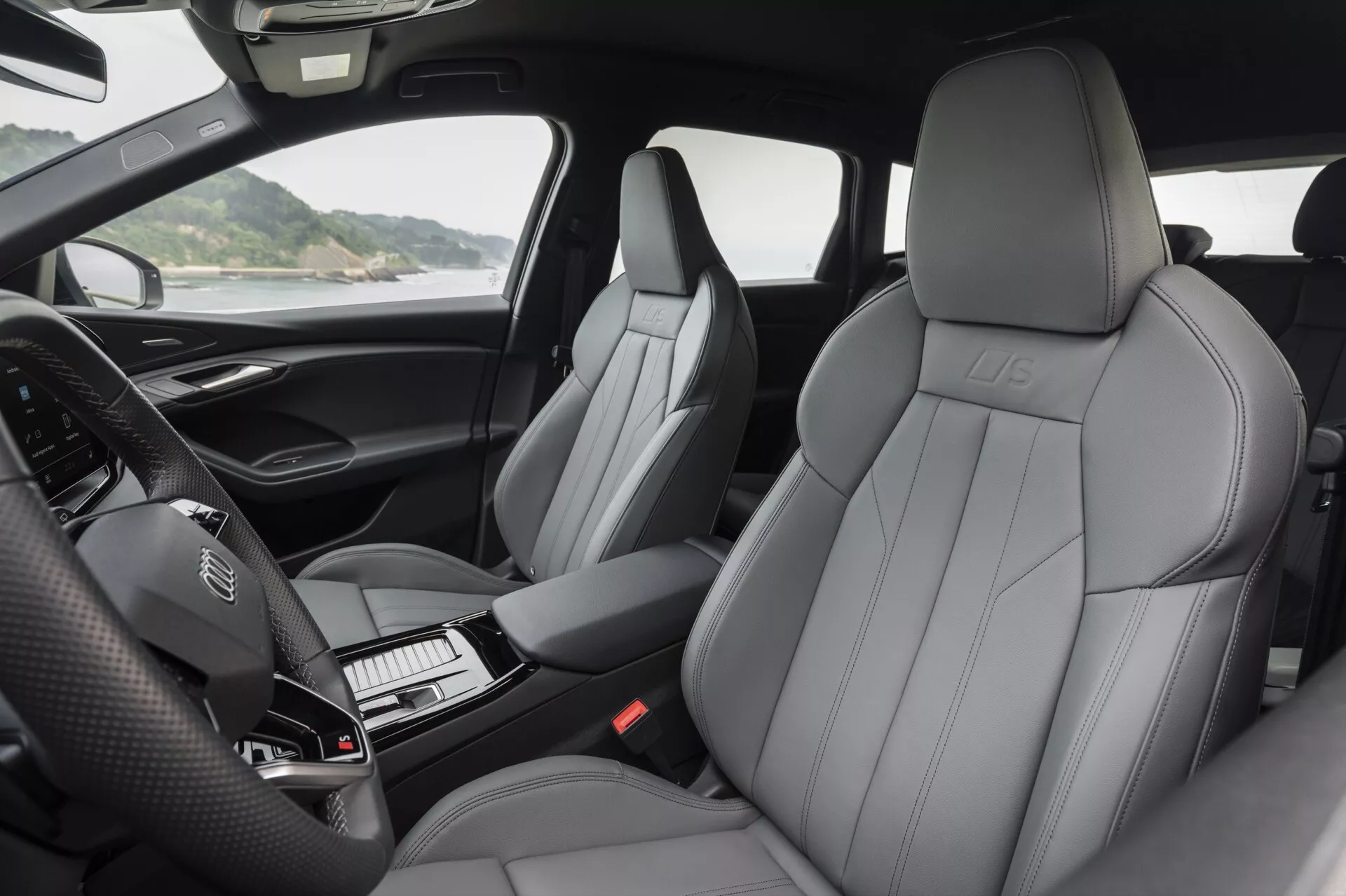 Pohled na interiér Audi Q6 e-Tron 2024, který ukazuje luxusní kožená přední sedadla s výrazným šitím, embosovaným logem a moderním designem. Fotografie zdůrazňuje palubní desku s volantem s integrovanými ovládacími prvky a nezapnutým digitálním přístrojovým štítem. Částečně je vidět středová konzola s řadicí pákou a uspořádanými tlačítky. Obrázek zachycuje čalouněné opěrky dveří s ovládáním pro zrcátka, okna a možná nastavení sedadel. Přirozené světlo zdůrazňuje komfort a vysokou kvalitu materiálů v kabině.