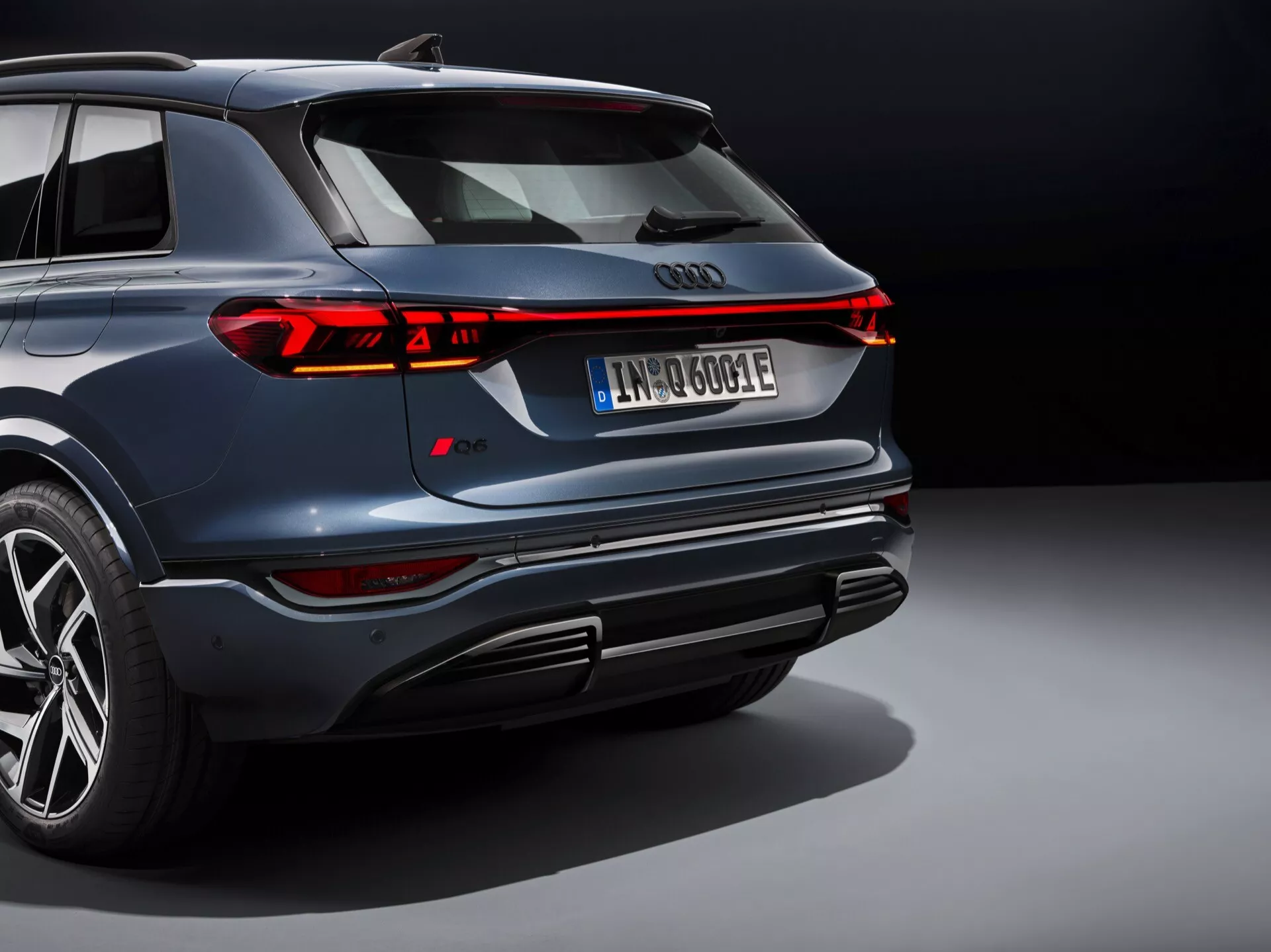 Tento obrázek zobrazuje moderní SUV, pravděpodobně Audi Q6 e-tron 2024, z pohledu shora zezadu. Ikonické čtyři kruhy Audi jsou výrazně umístěny vzadu, obklopené elegantním nepřerušovaným LED zadním světlem, které se táhne po celé šířce vozu, s segmentovými brzdovými světly a směrovkami. Tmavě námořní modrý vůz se může pochlubit leštěným kovovým povrchem, odvážným designem nárazníku s integrovanými výfuky a difuzorem, vícepaprskovými slitinovými koly s nízkoprofilovými pneumatikami a registrační značkou naznačující elektrický nebo hybridní pohon. Profesionální osvětlení a neutrální pozadí zdůrazňují luxusní a pokročilý design vozu, zdůrazňují jeho sportovní a vysoce výkonné atributy.