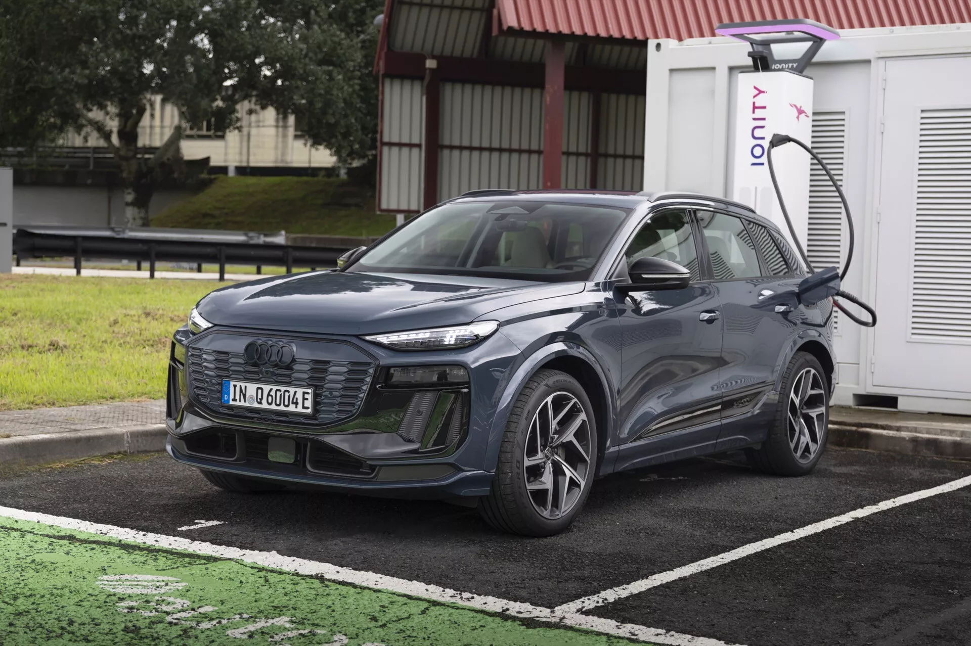 Obrázek ukazuje moderní SUV Audi Q6 e-Tron 2024 zaparkované u nabíjecí stanice pro elektrovozy. Auto je zobrazeno z lehce šikmého předního pohledu, zvýrazňující jeho mřížku, světlomety a část kapoty a čelního skla. Je umístěno diagonálně s přední částí mírně otočenou doleva. Viditelná je levá strana vozidla včetně předního kola na straně řidiče, dveří a bočního zrcátka. SUV je připojeno k nabíjecí stanici na levé přední straně nad kolem. Auto má elegantní linie, uzavřenou mřížku typickou pro elektrovozy a pokročilé LED světlomety. Detaily naznačují možnost pokročilých asistenčních systémů pro řidiče, jako je adaptivní tempomat, asistent udržování jízdního pruhu a systém prevence kolizí. Obrázek je jasný a čistý, pořízený během dne bez pohybové neostrosti, takže jsou všechny designové a technologické prvky snadno viditelné.