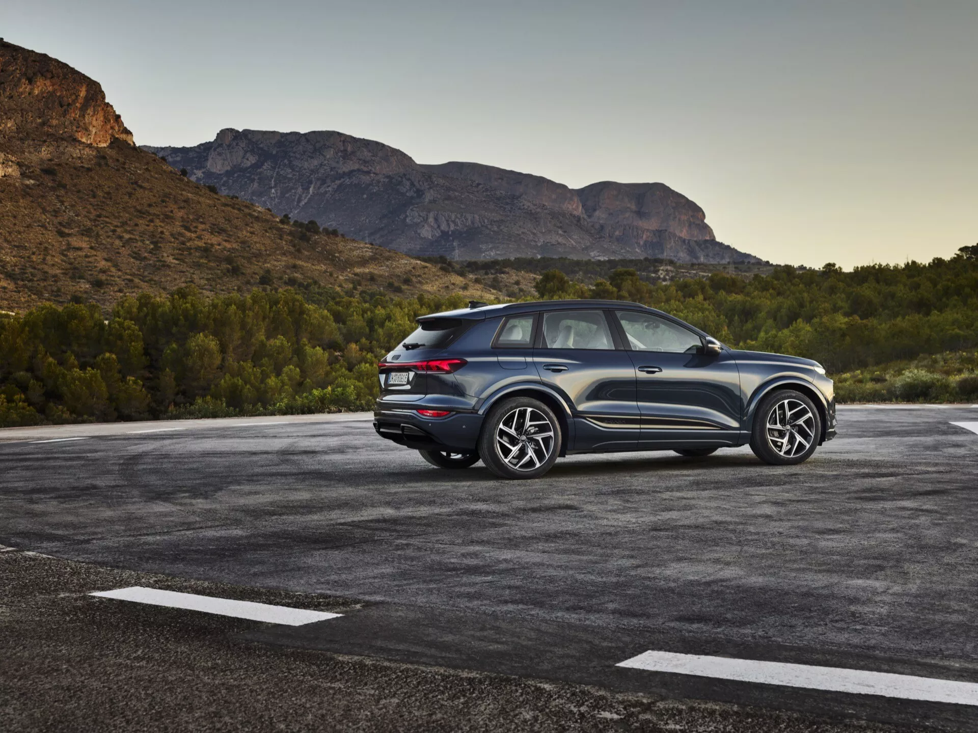 Obrázek SUV Audi Q6-e-tron 2024 zaparkovaného na venkovním parkovišti s přírodní, možná horskou krajinou v pozadí. Fotografie zachycuje tříčtvrteční zadní pohled, zdůrazňující boční i zadní designové prvky. Strana řidiče je zřetelně viditelná, předvádějící designové linie, zadní okna a design kol. Skloněná střecha naznačuje sportovní, kupé-SUV styl. Moderní LED zadní světla, viditelné senzory nebo odrazky v zadním nárazníku a dvoubarevné schéma zdůrazňují výšku a postoj. Velká hliníková kola a výrazné pneumatiky naznačují vyšší výbavový stupeň se sportovním vzhledem. Vozidlo se zdá být blízko kameře, poskytující detailní pohled. Významné prvky zahrnují LED osvětlení, parkovací senzory, vysoce výkonná kola a pneumatiky, aerodynamický design a možná pokročilé asistenční systémy pro řízení a pohon všech kol.