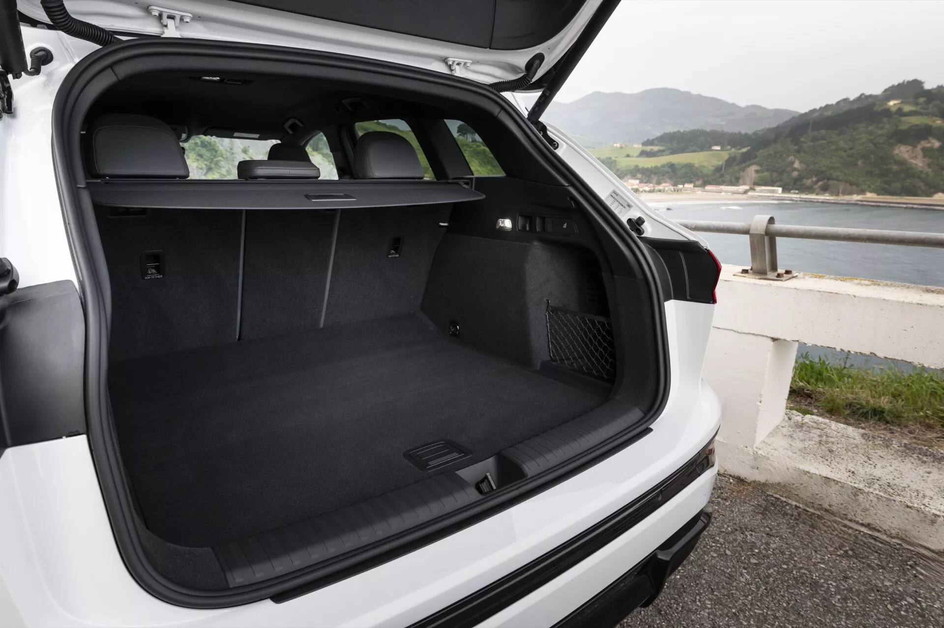 Obrázek zobrazuje nákladní prostor bílého Audi Q6 e-Tron 2024 hatchbacku nebo SUV, pohled zezadu s otevřeným víkem kufru a mírným pohledem dolů. Kufr s černým kobercem je prázdný, což ukazuje jeho prostornost a čistotu. Zadní sedadla jsou vzpřímená, což naznačuje možnost přepravy cestujících nebo sklopení sedadel pro větší nákladový prostor. Zakrytí nákladu je umístěno těsně za zadními sedadly, ale není roztaženo, což umožňuje plný pohled do kufru. Na levé straně nákladního prostoru je 12voltová zásuvka a na pravé straně se zdá být odkládací prostor pro uchování drobných předmětů. Viditelná je zadní stěrač. Vozidlo je zaparkováno u pobřeží, což naznačuje přímořskou lokaci. V kufru nejsou žádné předměty ani lidé.