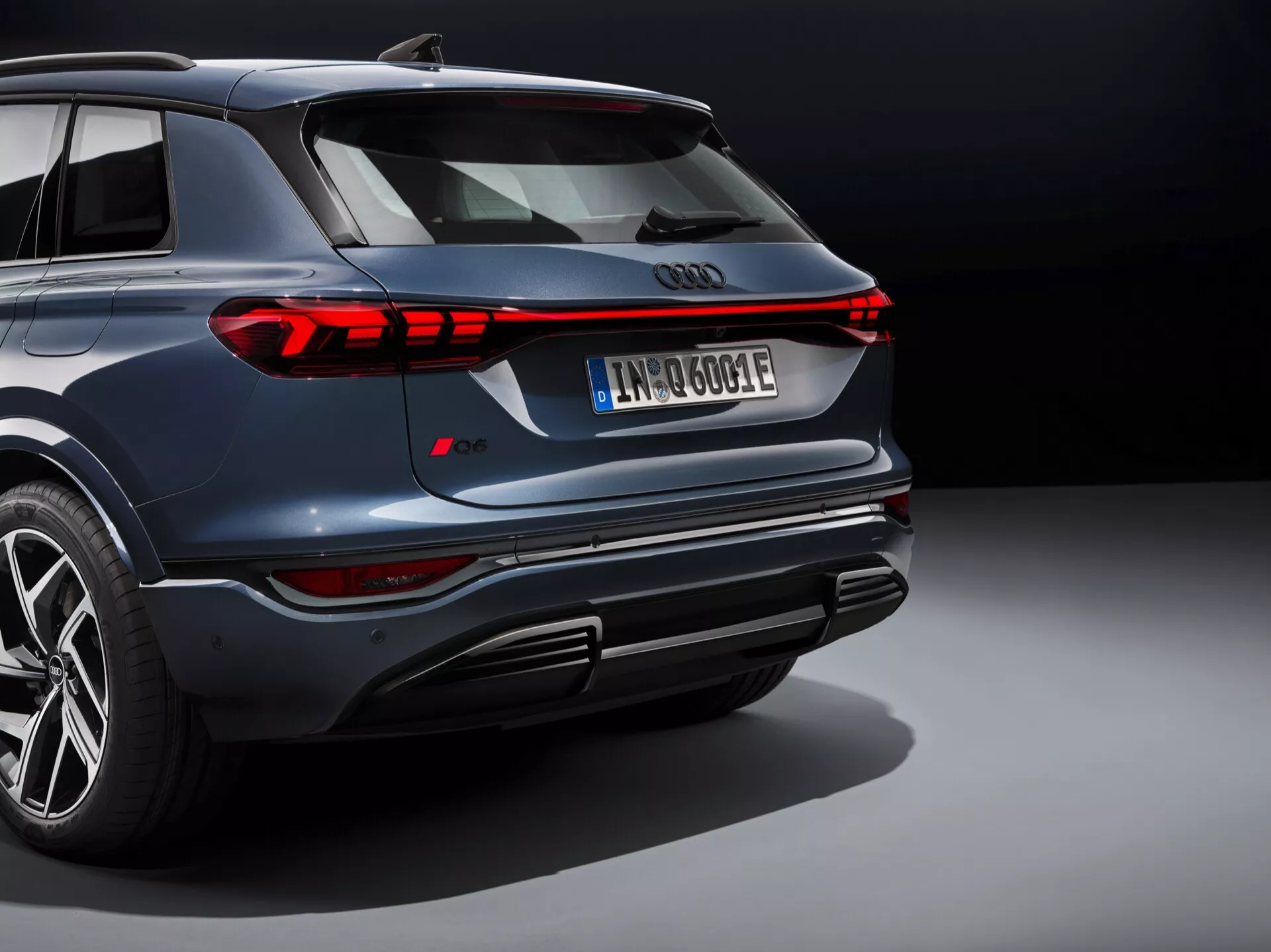 Zadní úhel SUV Audi Q6 e-tron 2024 s metalickou modrou barvou a moderními LED zadními světly. Obrázek zdůrazňuje elegantní design vozidla nízkým úhlem pohledu, který zdůrazňuje zadní nárazník, integrované zakončení výfuků, difuzor a velká litá kola. Výrazný horizontální světelný podpis a nápadný střešní spoiler podporují sportovní estetiku. Evropská registrační značka naznačující elektrický nebo hybridní pohonný systém s viditelným vzorkem pneumatik naznačující výkonné pneumatiky. Foceno ve studiovém nastavení s kontrolovaným osvětlením.
