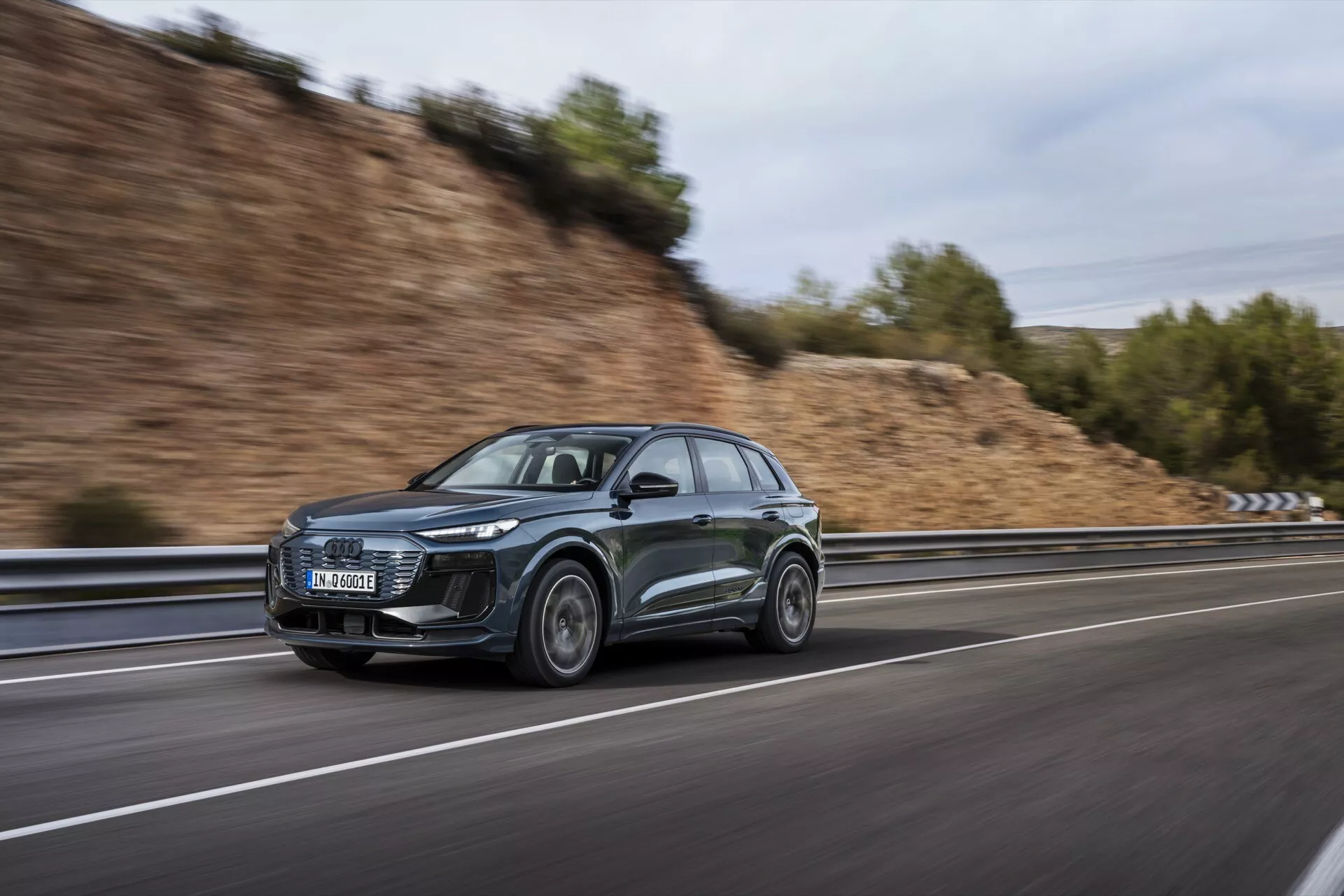 Tento obrázek zachycuje moderní a sofistikované Audi Q6 e-tron 2024 SUV, fotografované z bočního úhlu při pohybu. Moderní design vozidla je zvýrazněn výraznými LED světlomety a aerodynamickými prvky, včetně šikmé střechy a aerodynamických bočních zrcátek. Auto, dokončené v tmavé barvě, pravděpodobně šedé nebo černé, jede po rychlostní silnici, což je patrné z rozmazaného pozadí vytvořeného pomocí techniky panningu. Přední kolo je lehce natočeno směrem ke kameře, což ukazuje design litých kol. Další viditelné prvky zahrnují možnou integrovanou technologii ve světlometech a bočních zrcátkách, stejně jako možný užitný zaměření na střechu.